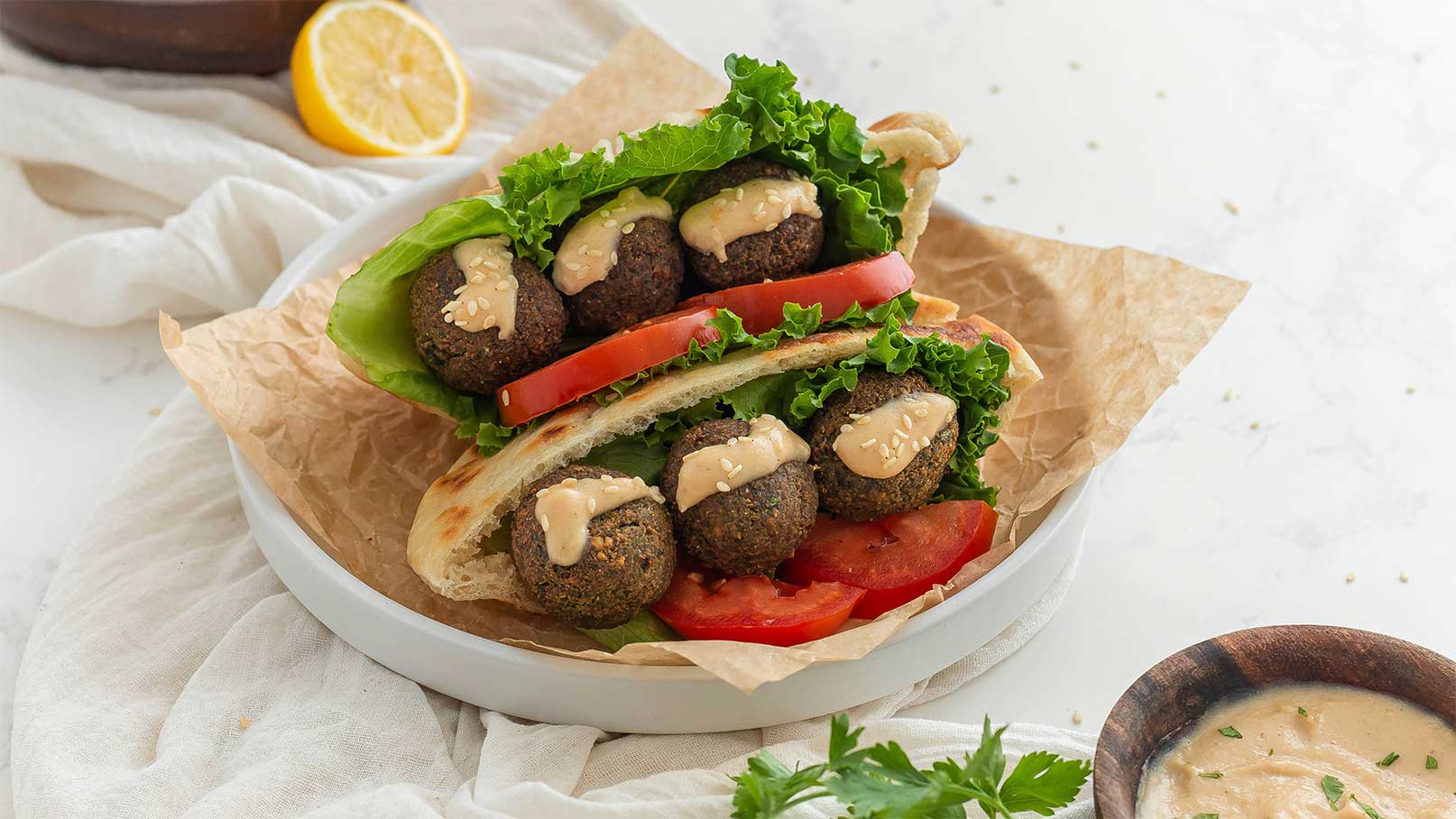 Ein Teller mit einem Fladenbrot gefüllt mit Falafel, frischem Salat und Tomatenscheiben. Die Falafel sind mit einer Creme, vermutlich aus Sesam, garniert. Das Gericht ist auf braunem Papier angerichtet und hebt eine gesunde, pflanzliche Mahlzeit hervor.