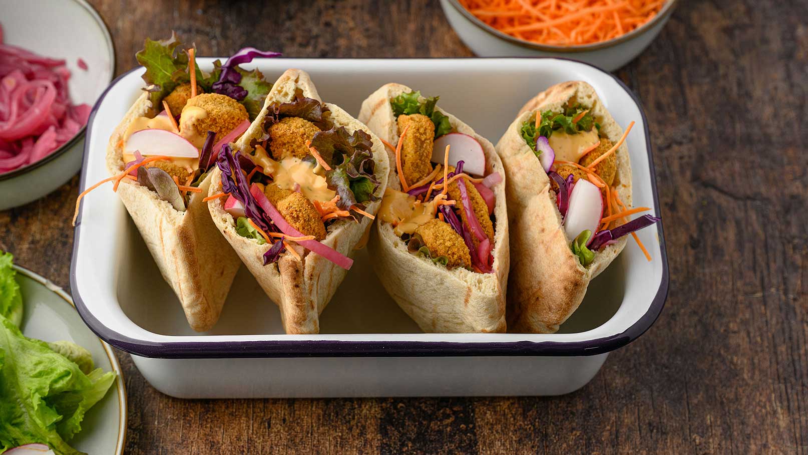 Drei gefüllte Pita-Brote in einer Schüssel, die mit frischem Salat, knusprigen Bällchen, buntem Gemüse und einer cremigen Sauce angerichtet sind. Im Hintergrund sind zusätzliche Zutaten wie geriebene Karotten sichtbar.