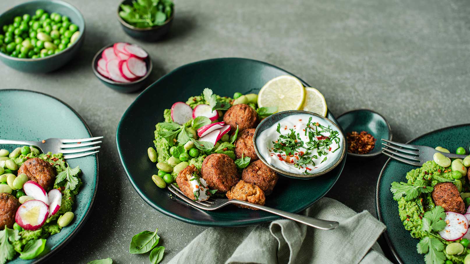 Eine grüne Schüssel mit einer bunten Mischung aus Falafel, Erbsen, Radieschen und frischen Kräutern. Daneben steht eine kleine Schüsseln mit Joghurt und frischen Kräutern. Limettenscheiben ergänzen das Gericht. Die Anrichtung ist appetitlich und farbenfroh.