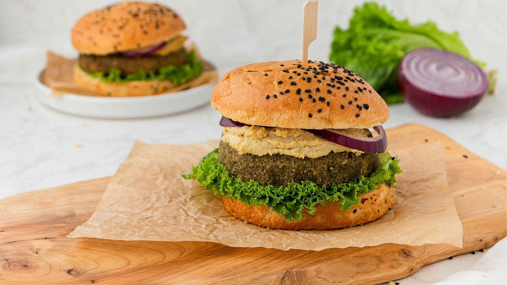Ein üppiger Burger mit einem bunten pflanzlichen Patty, belegt mit frischem Blattgrün, roten Zwiebeln und einer cremigen Sauce. Der Brötchenboden ist knusprig, während das Oberteil mit schwarzen Sesamsamen bestreut ist. Im Hintergrund sind weitere Zutaten zu sehen.