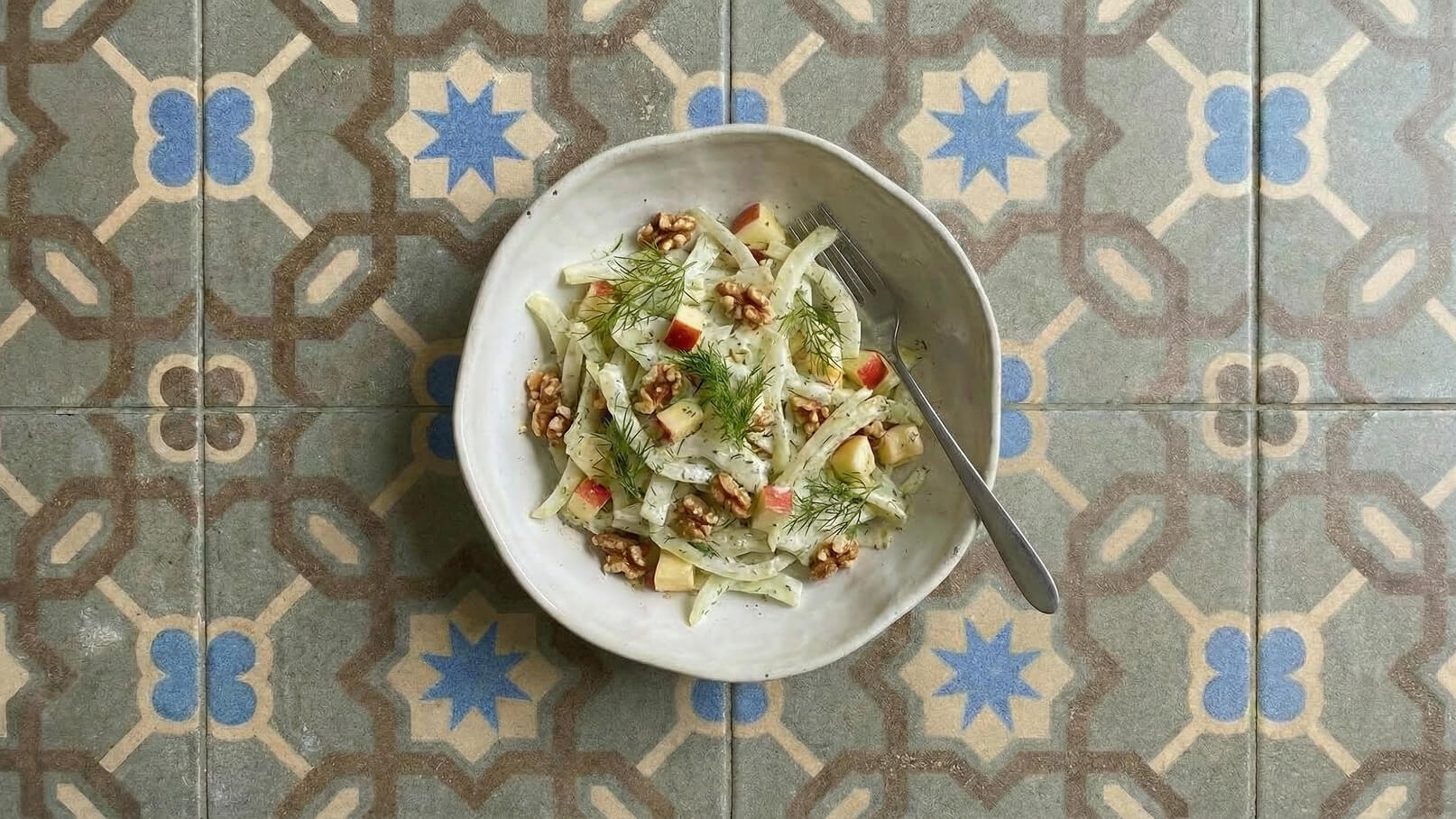 Ein tiefes, weißes Schälchen enthält einen frischen Salat aus Fennel, Äpfeln und Walnüssen, garniert mit frischem Dill. Eine Gabel liegt neben dem Schälchen. Der Hintergrund zeigt ein kunstvolles Fliesenmuster in gedeckten Farben.