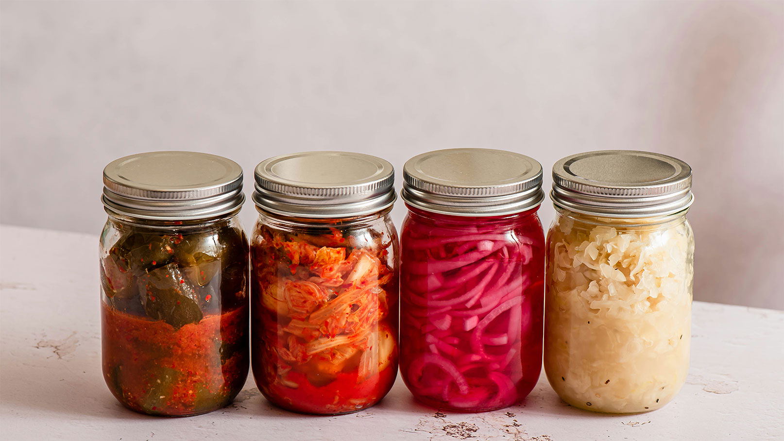 Vier Einmachgläser stehen nebeneinander, gefüllt mit verschiedenen eingelegten Gemüsevariationen. Von links nach rechts sind sie mit grünen und roten Gemüsen, Kimchi und roten Zwiebeln sowie einem Glas mit weißem Sauerkraut gefüllt. Alle Gläser haben einen silbernen Schraubdeckel.