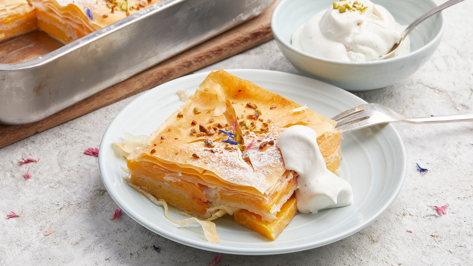 Ein Stück Baklava auf einem hellen Teller, garniert mit gehackten Pistazien und Puderzucker. Neben dem Baklava befindet sich ein Klecks Sahne. Im Hintergrund sieht man eine metallene Backform mit mehr Baklava.