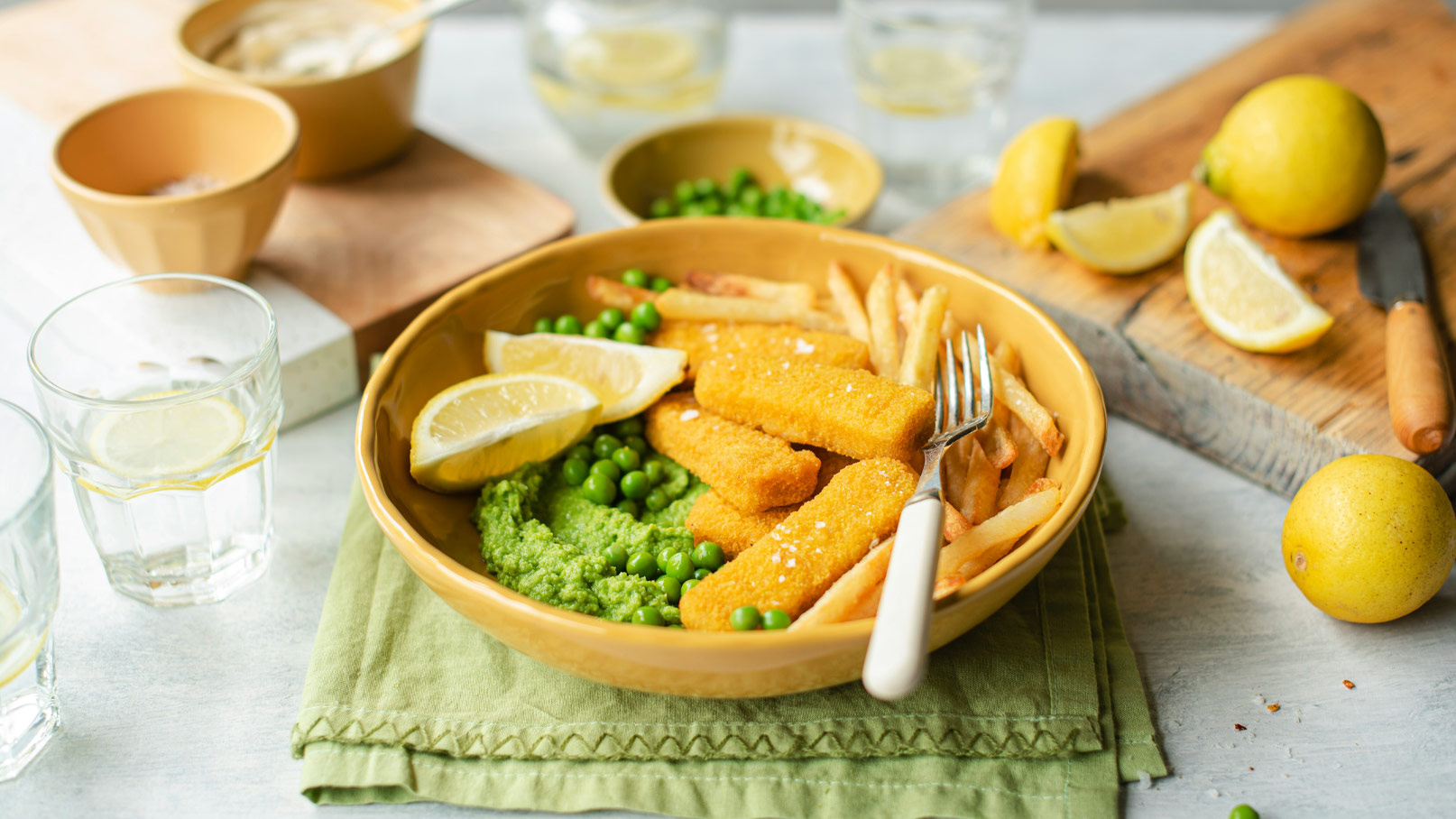 Eine Schüssel mit paniertem Fisch, serviert mit Pommes frites, grünen Erbsen und einem grünen Püree. Zwei Zitronenscheiben liegen auf dem Fisch, und im Hintergrund sind weitere Zitrone und Wassergläser sichtbar. Das Ganze ist auf einem grünen Tuch angerichtet.