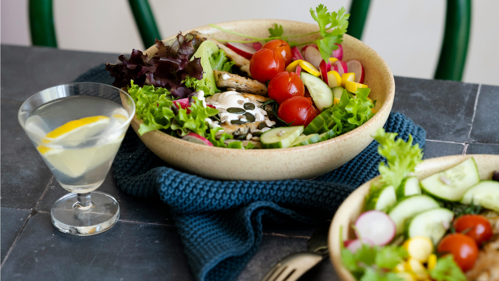 Eine Schüssel mit frisch zubereitetem Salat, gefüllt mit verschiedenen Blattsalaten, Kirschtomaten, Gurken, Radieschen und einem Dressing. Neben der Schüssel steht ein Glas mit klarem Getränk und einer Zitronenscheibe. Der Hintergrund ist unauffällig.