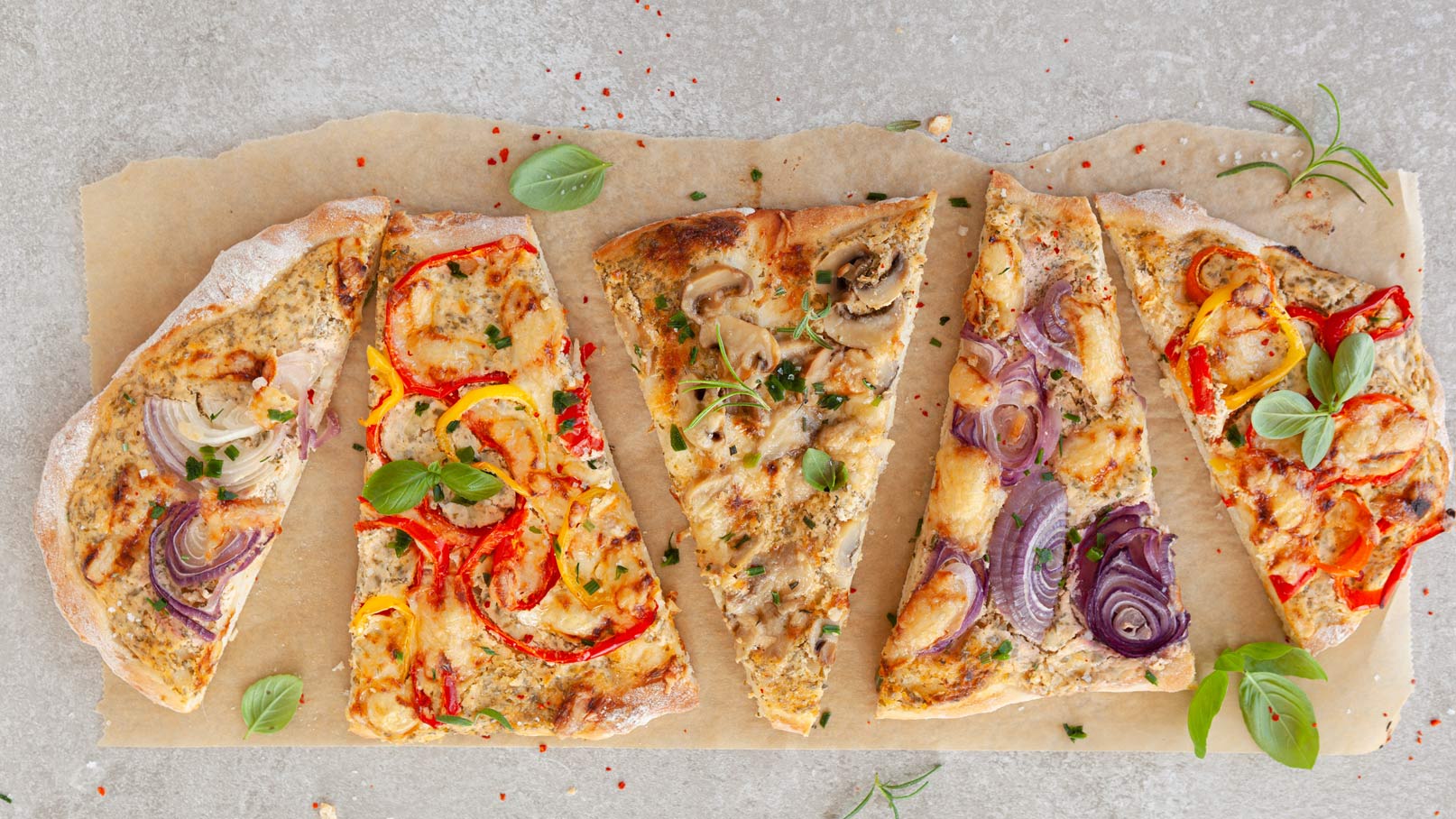 Fünf Stücke Pizza liegen auf Backpapier. Jedes Stück hat unterschiedliche Beläge: bunte Paprika, Zwiebeln, Pilze und Kräuter sind sichtbar. Die Pizza hat einen goldbraunen Rand und ist frisch belegt.