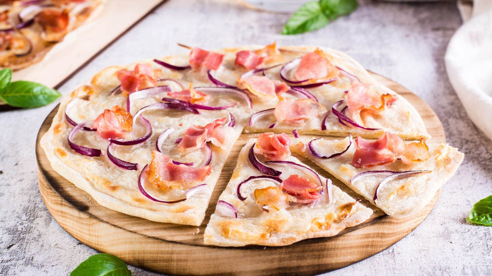 Ein frisch gebackener Flammkuchen auf einem Holzbrett, belegt mit roten Zwiebeln und gebratenem Speck. Der Flammkuchen ist in Stücke geschnitten und zeigt eine knusprige, goldene Kruste. Im Hintergrund sind einige grüne Basilikumblätter sichtbar.