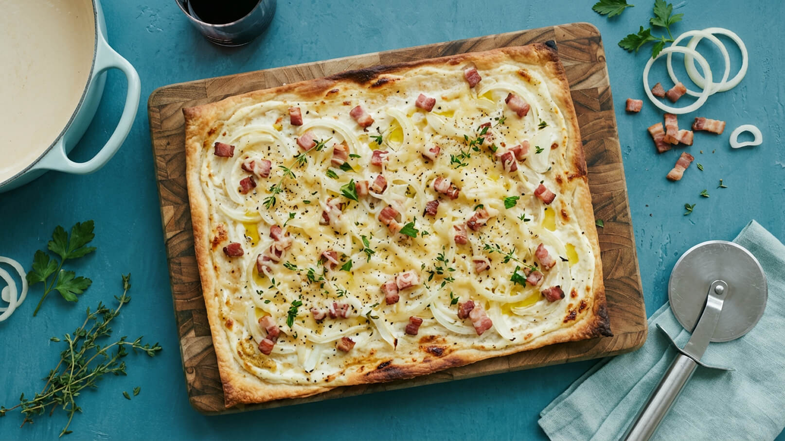 Ein frisch gebackener Flammkuchen liegt auf einem Holzbrett. Er ist belegt mit einer cremigen Schicht, Zwiebeln und kleinen Stückchen Speck. Frische Kräuter und Pfeffer sind verteilt, um den Geschmack zu verfeinern. Im Hintergrund sind einige Zutaten und ein Pizzaschneider sichtbar.