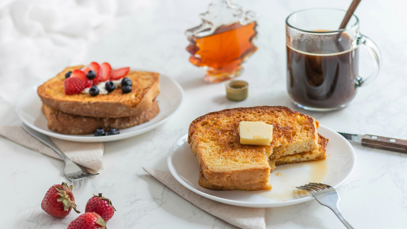 Ein Teller mit zwei Scheiben French Toast, eine davon mit einem Stück Butter darauf. Neben dem Teller befindet sich eine Tasse schwarzer Kaffee und ein Glas Ahornsirup. Am Rand liegen frische Erdbeeren und eine Serviette.
