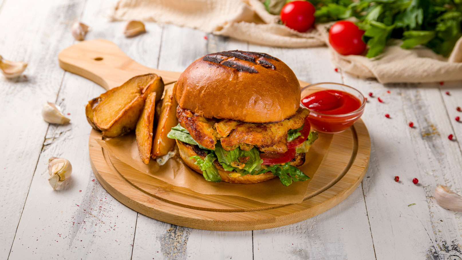 Ein frisch zubereiteter Burger mit gegrilltem Hähnchen, frischem Salat und Tomaten in einem weicheren Brötchen, serviert mit knusprigen Kartoffelspalten und einer kleinen Schüssel Ketchup auf einem Holzbrett.