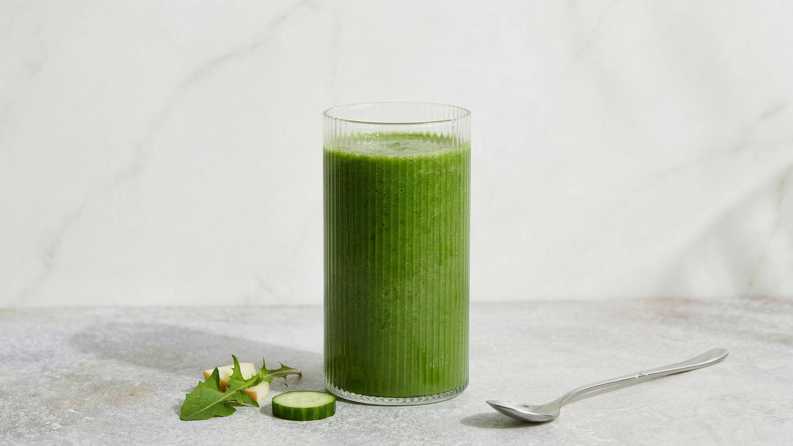 Ein Glas mit grünem Smoothie steht auf einer grauen Oberfläche. Neben dem Glas liegen ein paar frische Gurkenscheiben und ein Blatt. Ein Löffel befindet sich ebenfalls in der Nähe. Der Smoothie wirkt frisch und gesund.