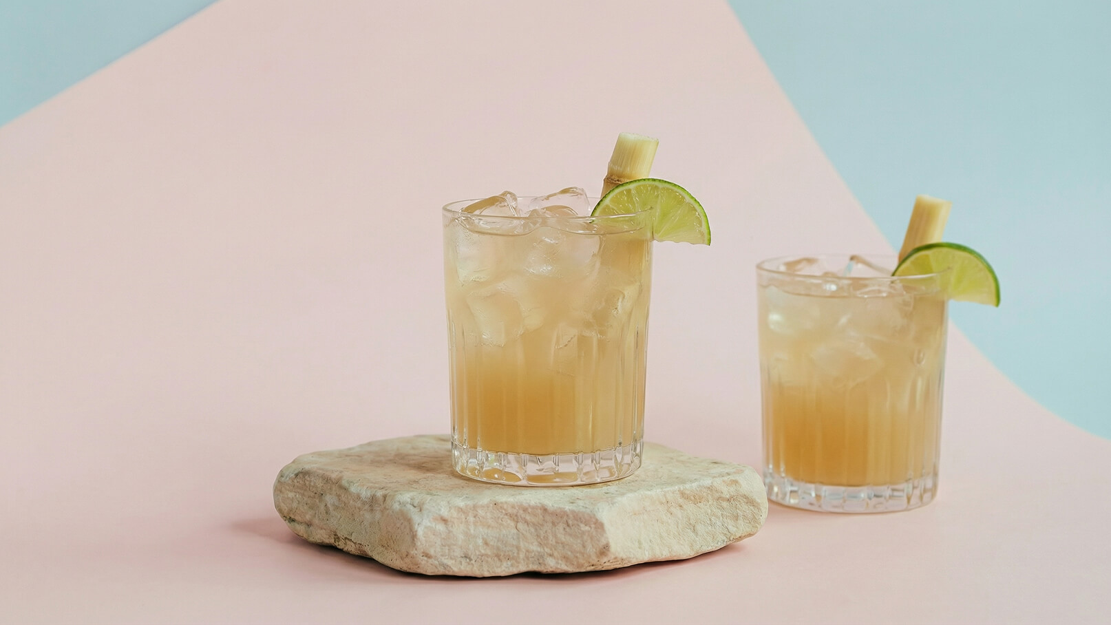 Zwei Gläser mit einem erfrischenden Getränk stehen auf einem hellen Stein. Die Gläser sind mit Eis gefüllt und mit Limettenscheiben sowie einem Stück Zuckerrohr dekoriert. Der Hintergrund ist in sanften, pastellfarbenen Tönen gehalten.