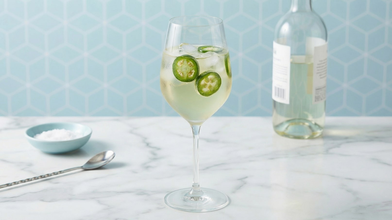 Ein elegantes Weinglas enthält einen Cocktail mit Eiswürfeln und dünn geschnittenen Jalapeños. Im Hintergrund steht eine Flasche Weißwein auf einer marmorierten Oberfläche. Ein kleiner, blauer Schüssel mit Salz ist ebenfalls sichtbar. Der Hintergrund hat ein geometrisches Muster in hellblau.
