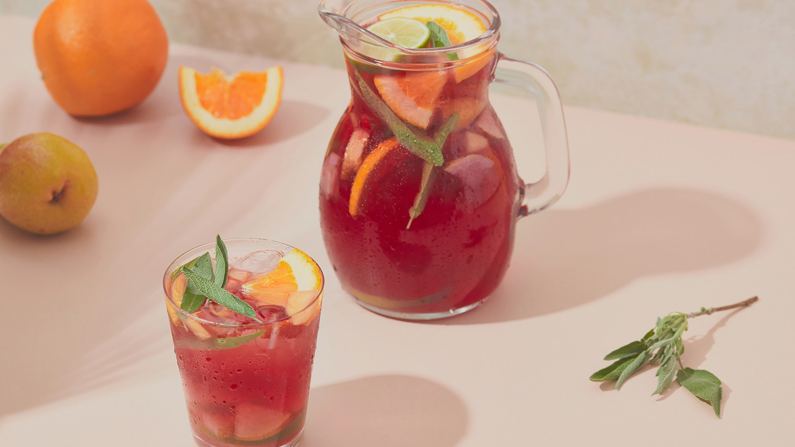 Ein Glas und eine Karaffe mit einem fruchtigen Getränk, angereichert mit frischen Orangenscheiben und Kräutern. Die Karaffe steht auf einem hellen Tisch, während eine Orange und ein Stück einer halben Orange daneben liegen.