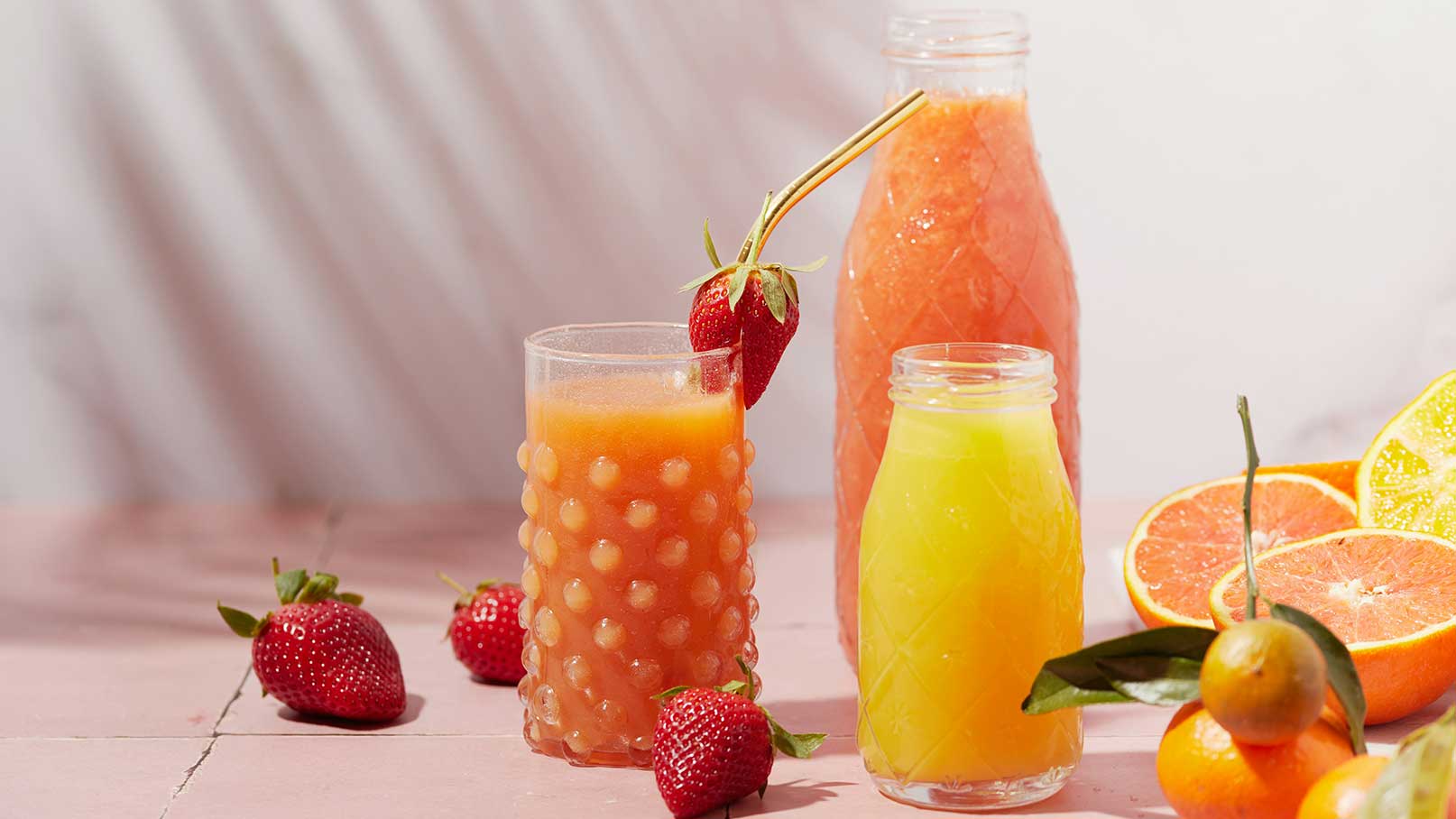 Zwei Gläser mit frischen Säften stehen auf einem Tisch, eines mit orangefarbenem Getränk und einem Strohhalm, das andere mit gelbem Saft. Neben den Gläsern liegen frische Erdbeeren und verschiedene Zitrusfrüchte, darunter Orangen und Zitronen. Das Bild strahlt Frische und Sommerlichkeit aus.