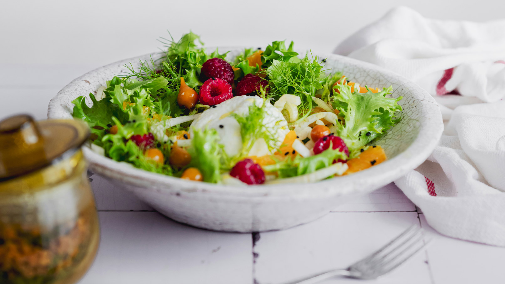 Ein frischer Salat in einer schüsselförmigen weißen Schale. Der Salat besteht aus verschiedenen grünen Blattsalaten, Himbeeren, gelben Beeren und feinen Gemüsestreifen. Im Hintergrund steht ein Glas mit einer unkenntlichen Zubereitung.