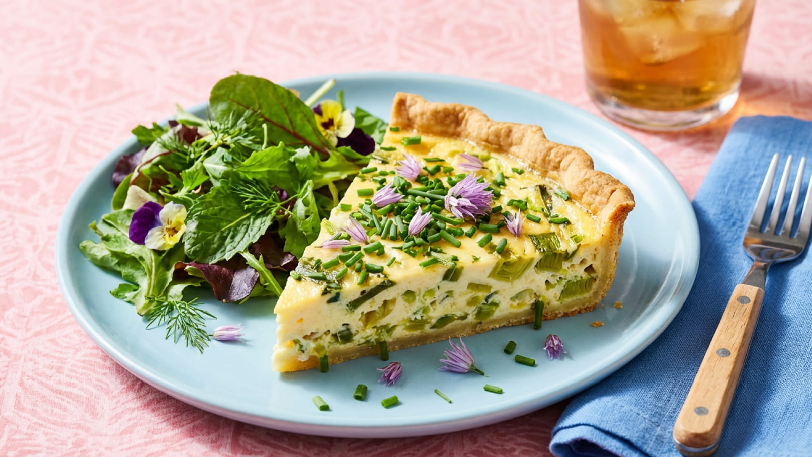 Ein Teller mit einer Scheibe Quiche, garniert mit Schnittlauch und Essblumen, liegt neben einem bunten Salat aus verschiedenen Blattsalaten. Im Hintergrund ist ein Glas mit einem Getränk zu sehen. Der Teller ist auf einem hellen Tischset platziert.