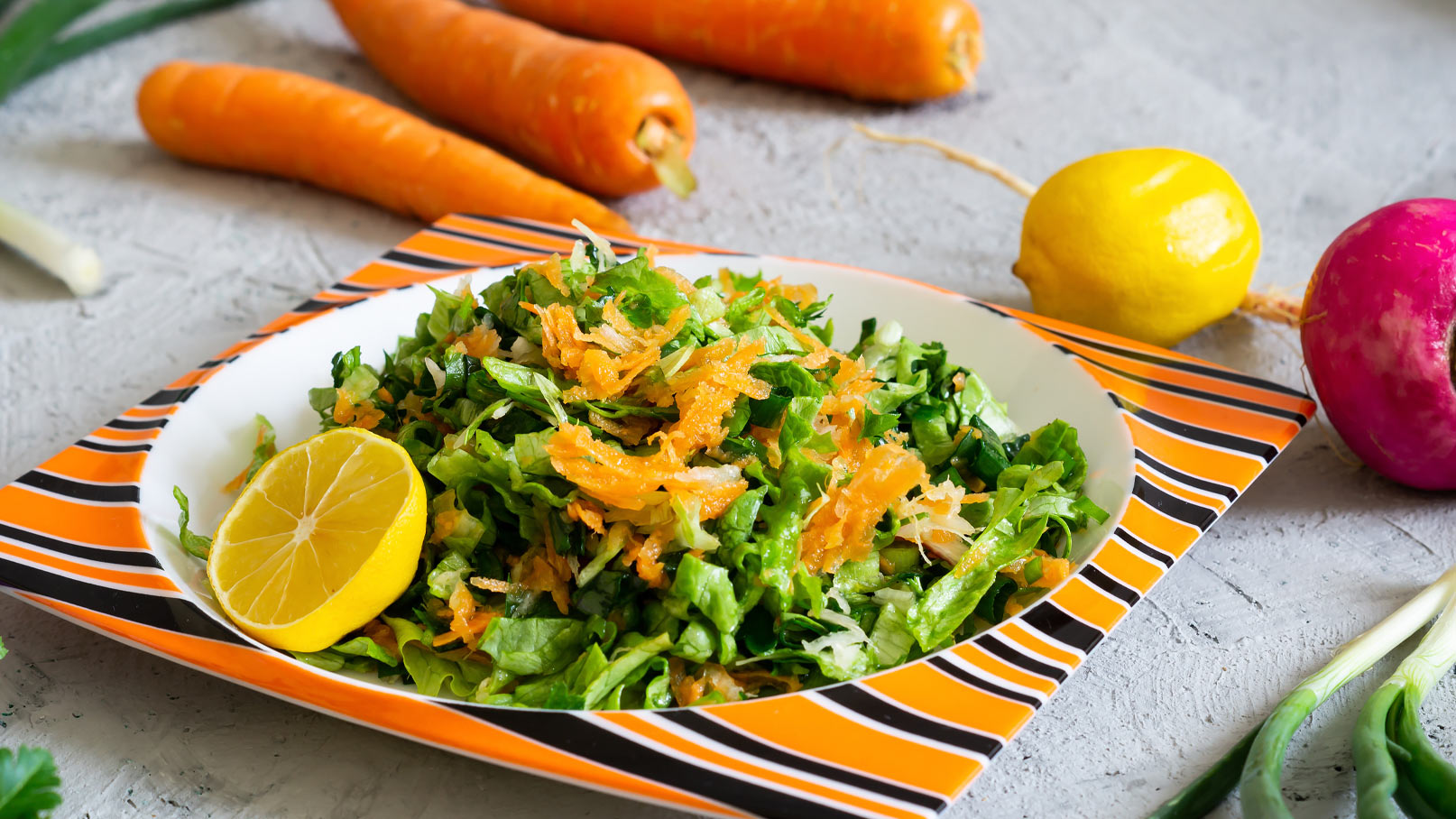 Ein frischer Salat aus grünen Blättern und geraspelten Karotten, angerichtet auf einem orange-gestreiften Teller. Eine Zitronenscheibe dekoriert das Gericht. Im Hintergrund sind weitere frische Zutaten wie Karotten und Zitronen sichtbar.