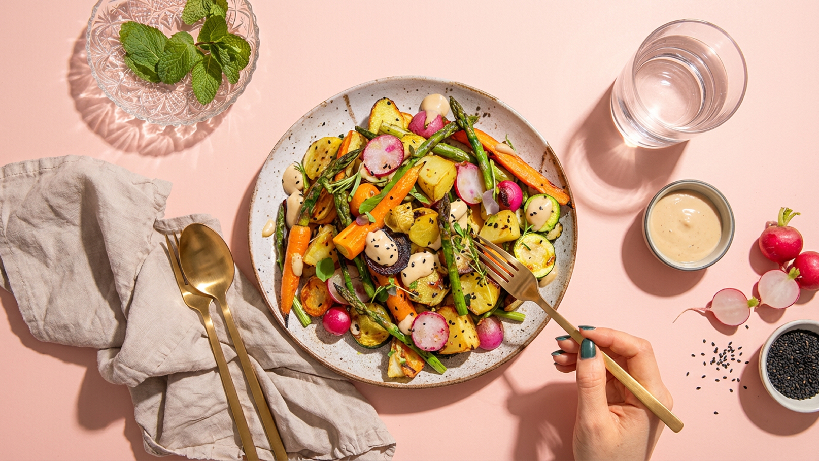 Ein Teller mit buntem, geröstetem Gemüse, darunter Karotten, Radieschen, Zucchini und grüne Bohnen, ist auf einem rosa Hintergrund angerichtet. Eine Hand hält eine goldene Gabel, die sich dem Gemüse nähert. Neben dem Teller befindet sich eine Schüssel mit einer Sauce und einige Radieschen.
