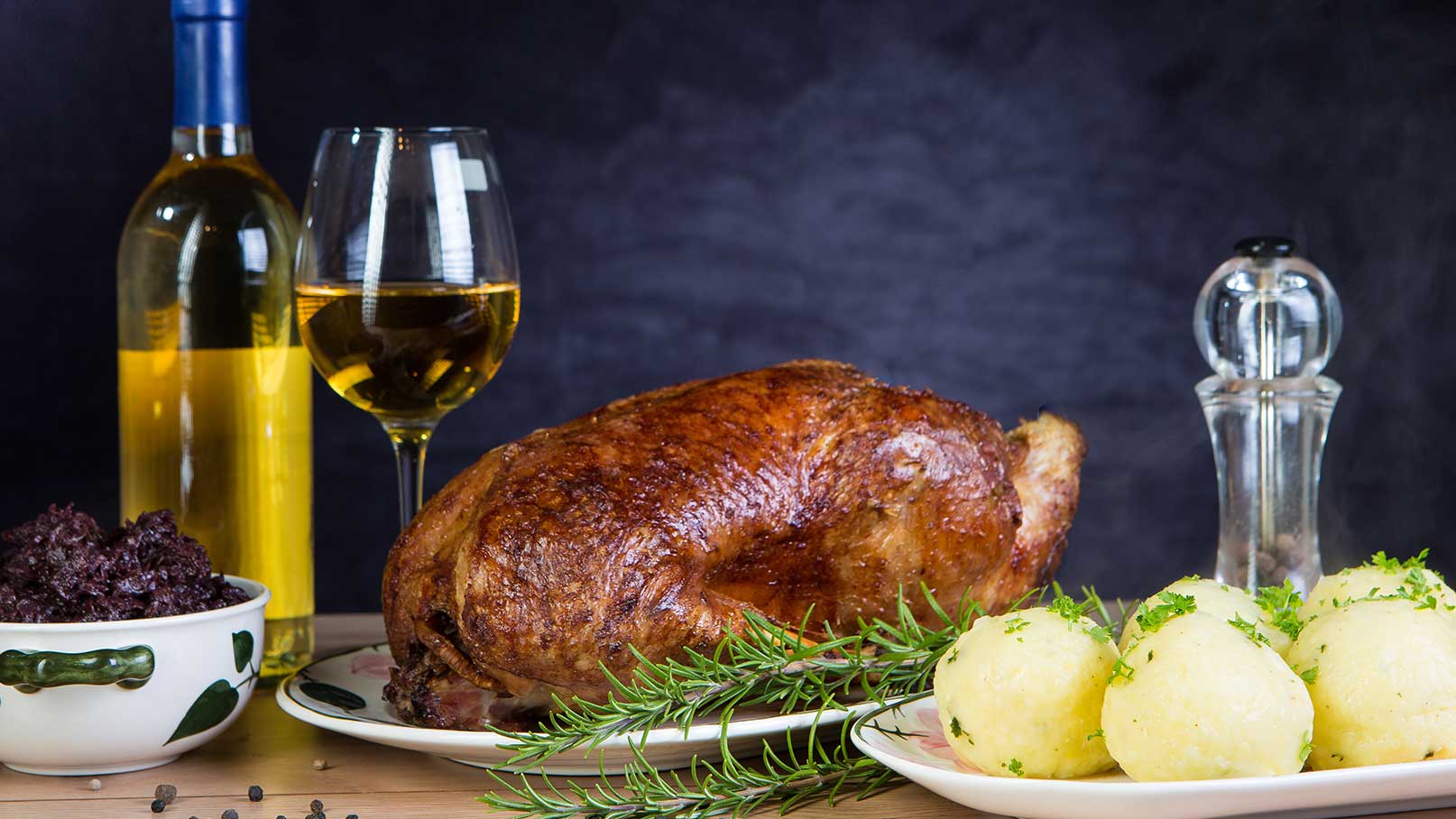 Ein gebratener Gans auf einem Teller, begleitet von zwei Portionen Kartoffelklößen und frischem Rosmarin. Im Hintergrund stehen zwei Weingläser mit Weißwein und eine Gewürzmühle. Die Szene vermittelt eine einladende, festliche Atmosphäre.