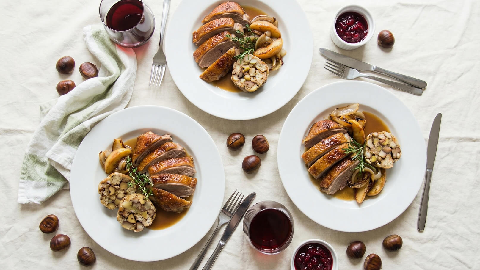 Drei Teller mit gebratener Entenbrust, garniert mit frischen Kräutern. Dazu gibt es geschmorter Apfel und ein Füllungsgericht. Neben den Tellern sind Kastanien und Gläser mit Rotwein sowie eine kleine Schüssel mit Preiselbeeren zu sehen. Der Hintergrund ist eine helle Tischdecke.