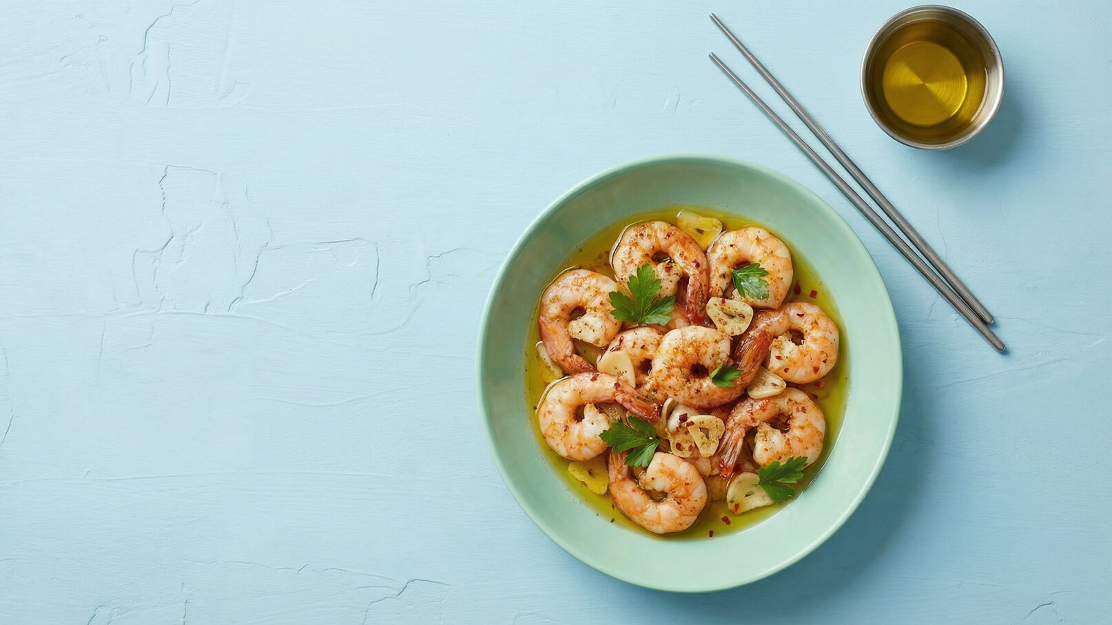 Ein grüner Teller mit gebratenen Garnelen, die in einer aromatischen Sauce schwimmen. Einige Scheiben Knoblauch und frische Petersilie sind sichtbar. Neben dem Teller liegen Essstäbchen und eine kleine Schüssel mit Öl. Der Hintergrund ist in sanften, hellblauen Tönen gehalten.