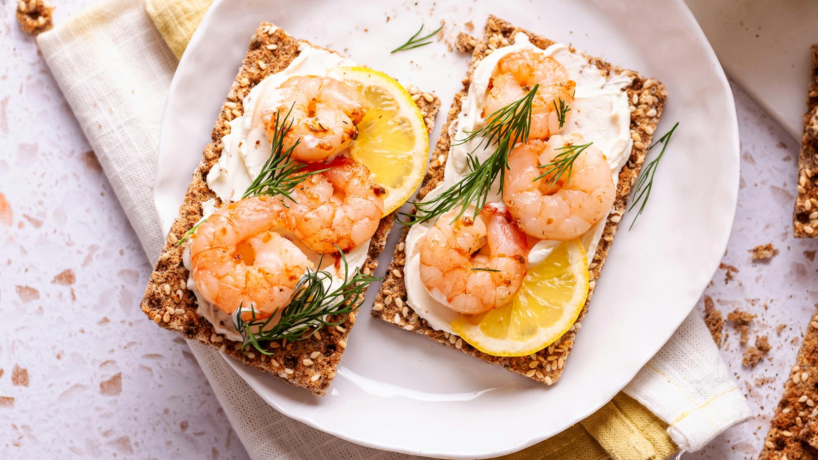 Ein Teller mit zwei knusprigen Brotstücken, belegt mit Sahne, garnierte Garnelen, Zitronenscheiben und frischem Dill. Der Teller befindet sich auf einer hellen Oberfläche, daneben liegt ein saftiges Tuch.