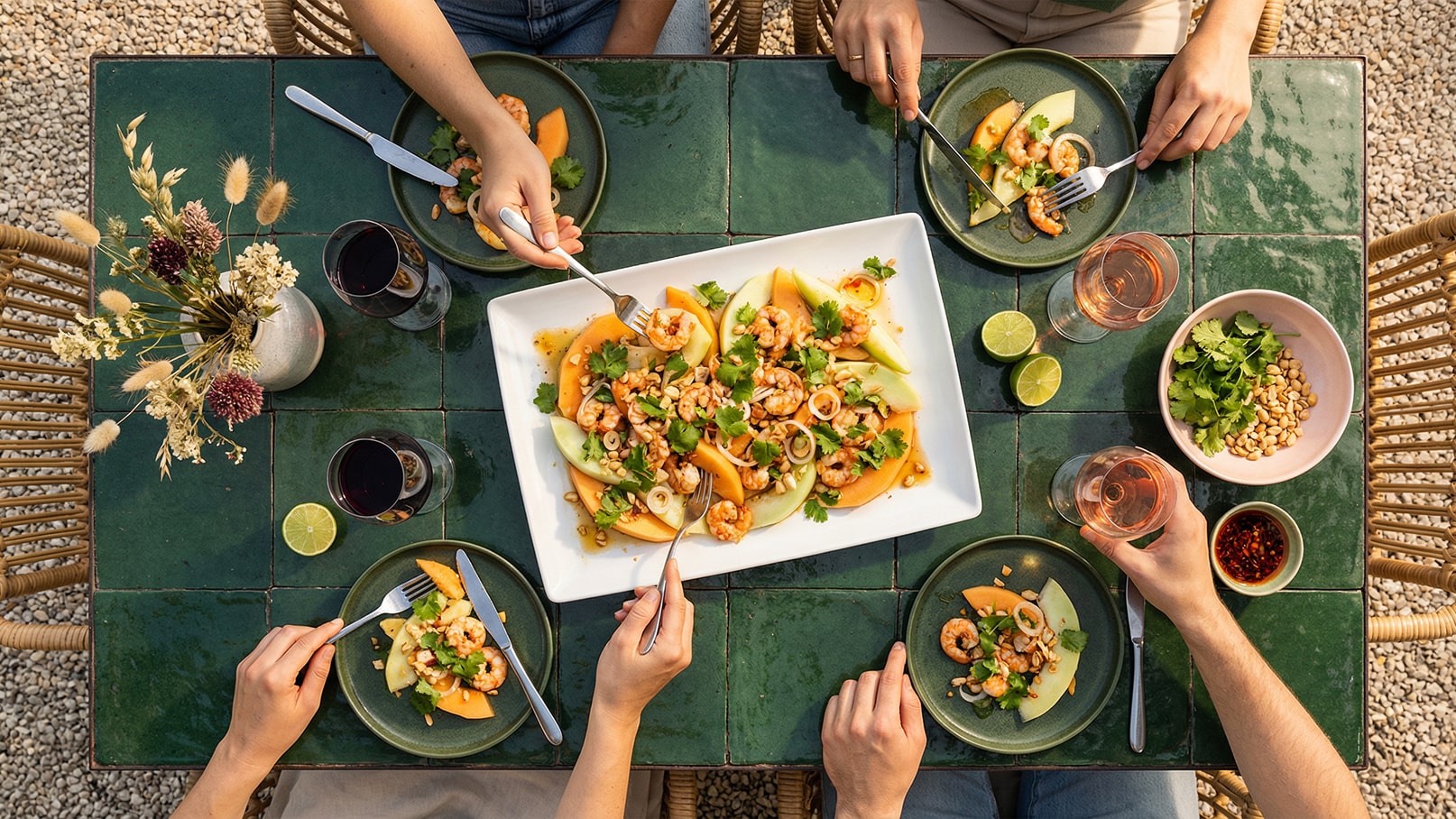 Ein gedeckter Tisch mit mehreren Personen, die ein buntes Gericht aus Garnelen, Melonen und Kräutern genießen. Auf dem Tisch stehen Weingläser, Limetten und Schalen mit Erdnüssen sowie frischem Koriander. Das Ambiente ist freundlich und einladend.