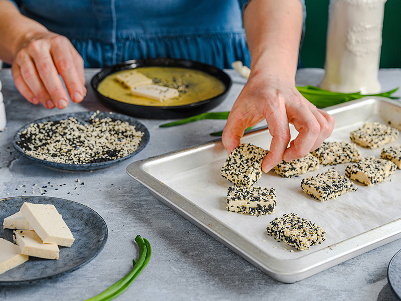 Eine Hand platziert ein Stück in Sesam gehüllten Tofu auf einem Backblech. Im Hintergrund sind weitere Tofu-Stücke und Schalen mit Zutaten wie schwarzem Sesam und einer Flüssigkeit sichtbar. Die Szene zeigt den Zubereitungsprozess eines Gerichts mit Tofu.