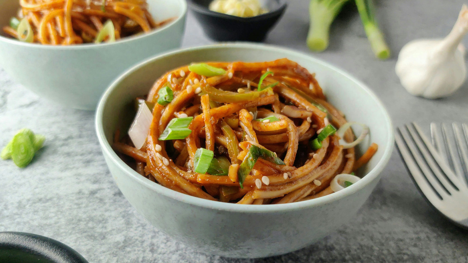 Eine Schüssel mit asiatischen Nudeln, garniert mit frischen Frühlingszwiebeln und Sesam. Im Hintergrund befindet sich eine zweite Schüssel Nudeln. Neben der Schüssel sind einige frische Knoblauchzehen und eine Gabel zu sehen.