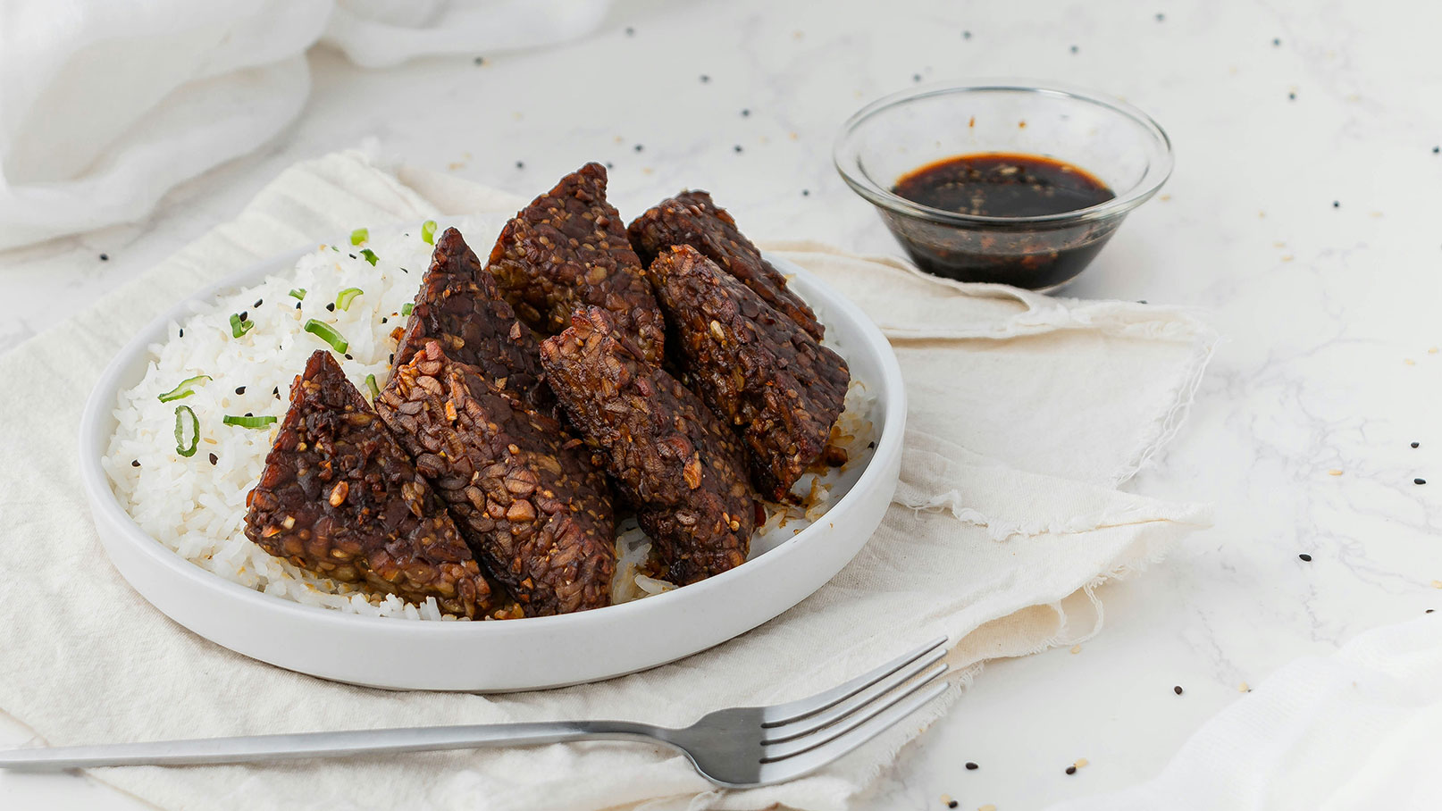 Eine weiße Schüssel enthält dampfenden, weißen Reis, garniert mit frischen Frühlingszwiebeln und schwarzem Sesam. Darauf liegen mehrere Stücke marinierter Tempeh, glänzend und knusprig. Neben der Schüssel befindet sich eine kleine Glas-Schale mit dunkler Soße und eine Gabel.