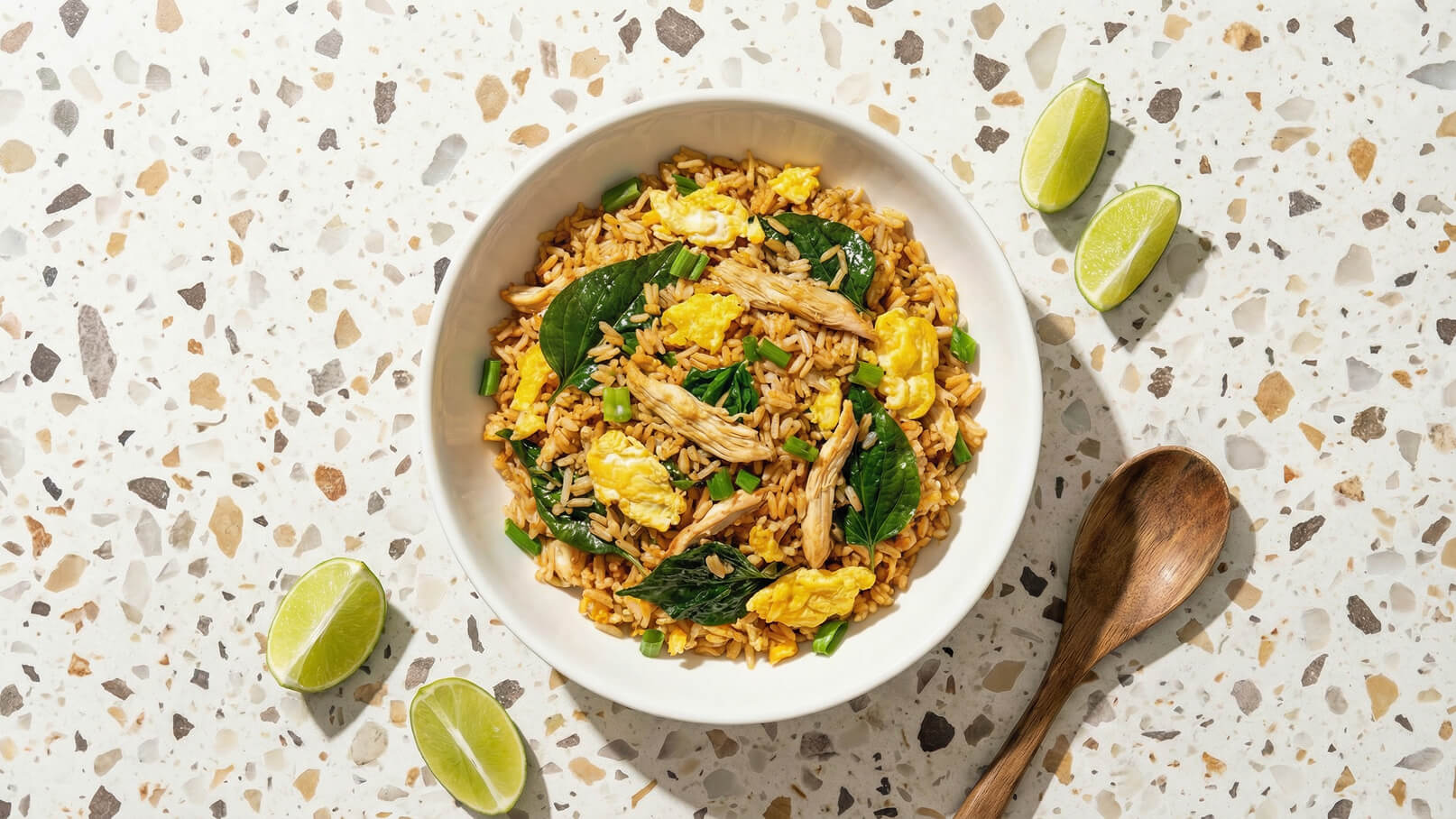 Eine Schüssel mit gebratenem Reis, garniert mit Spinat, Eiern und zerkleinertem Hähnchen. Neben der Schüssel liegen frische Limettenscheiben. Der Hintergrund besteht aus einer hellen, strukturierten Oberfläche.