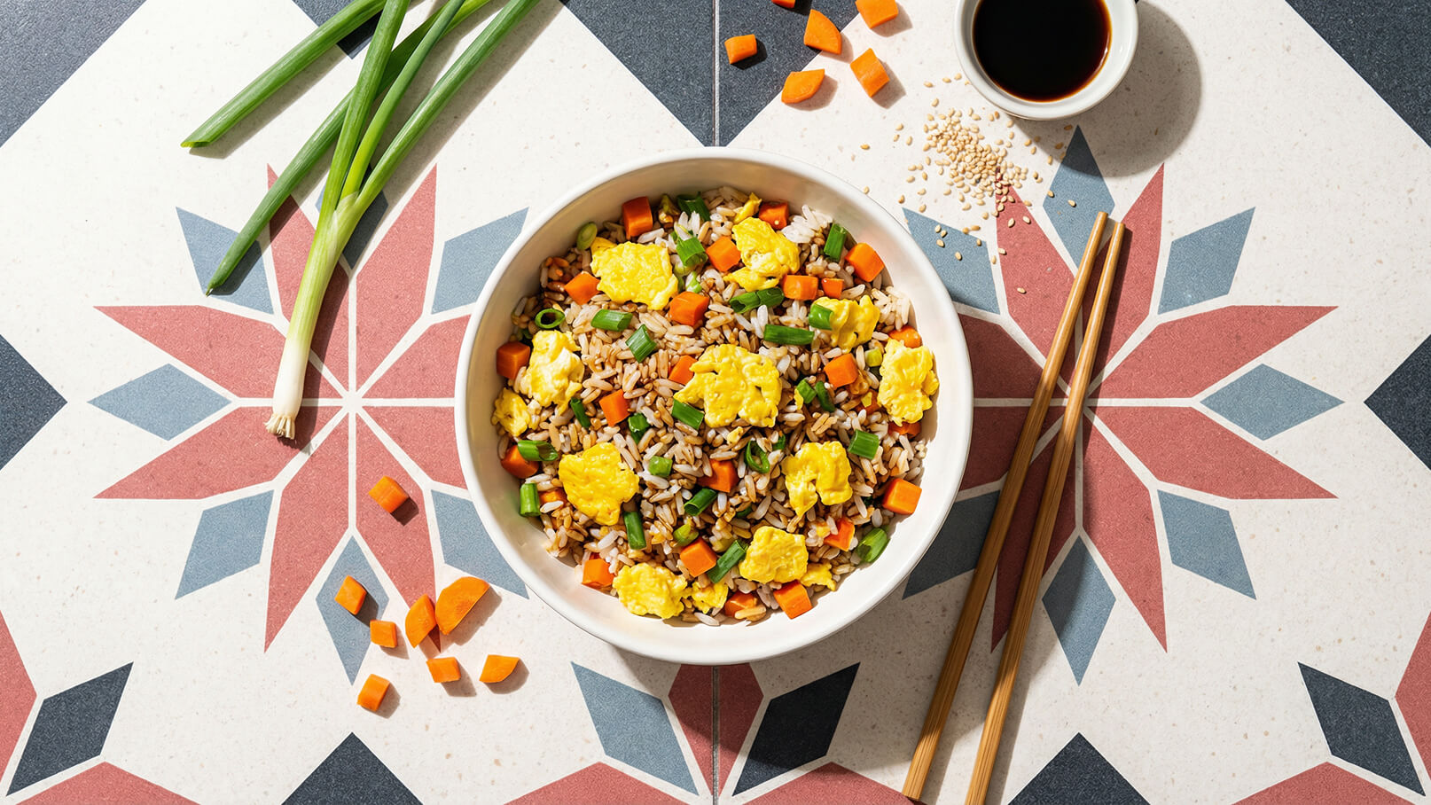 Eine weiße Schüssel mit gebratenem Reis, gemischt mit Karotten, grünen Bohnen und Rührei, auf einem gemusterten Tisch. Neben der Schüssel liegen Essstäbchen und eine kleine Schale mit Sojasauce. Einige frische Frühlingszwiebeln und Karottenstücke sind ebenfalls sichtbar.