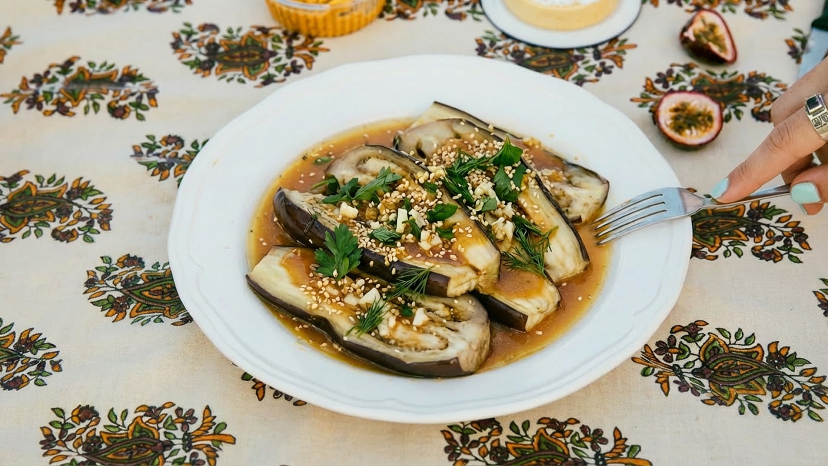 Ein Teller mit gegrillten Auberginenscheiben, übergossen mit einer dunklen Sauce. Die Auberginen sind mit frischen Kräutern, gehacktem Knoblauch und Sesam bestreut. Im Hintergrund sind ethnische Muster auf einem Tischcloth sichtbar. Eine Hand hält eine Gabel, die auf den Teller zeigt.