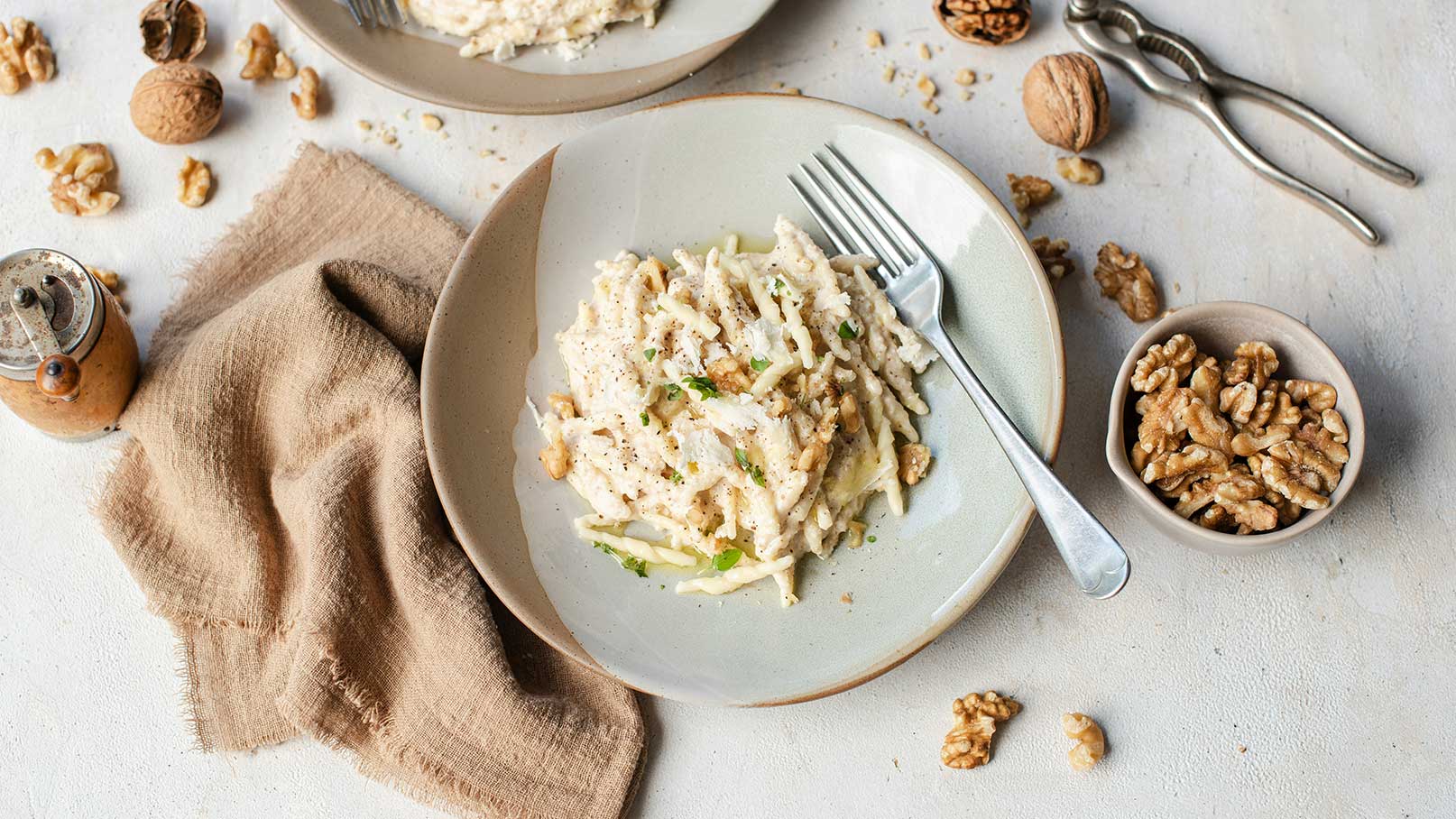 Ein Teller mit einem cremigen Salat aus geschnittenen Äpfeln und Walnüssen, garniert mit frischen Kräutern. Neben dem Teller liegt eine Gabel. Zusätzlich gibt es eine Schale mit weiteren Walnüssen und einige verstreute Nüsse auf dem Tisch.