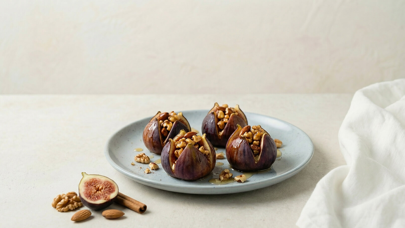 Auf einem hellen Teller liegen vier gefüllte Feigen. Sie sind mit Nüssen gefüllt, und ein wenig Honig ist darüber verteilt. Neben dem Teller liegen eine halbe Feige, einige Walnüsse, Mandeln und ein Zimtstängel. Die Präsentation ist ansprechend und einladend.