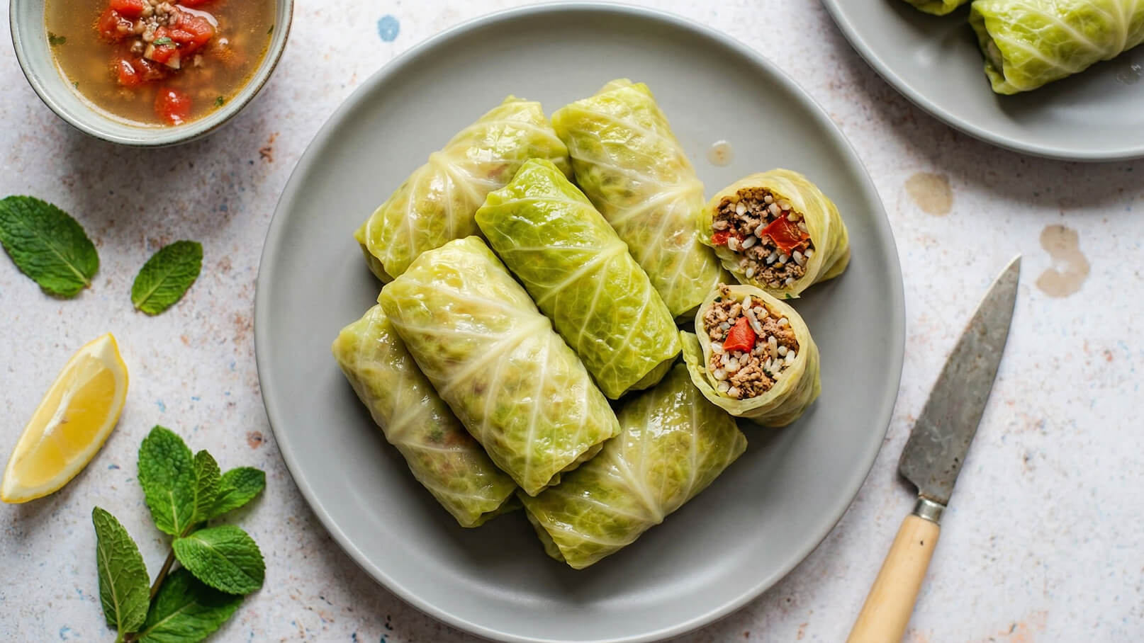 Auf einem grauen Teller liegen mehrere gefüllte Kohlrouladen. Eine der Rouladen ist aufgeschnitten, sodass die Füllung aus Reis, Fleisch und Paprika sichtbar ist. Daneben befindet sich ein kleiner Teller mit einer Brühe und kleine Stücke von roten Tomaten.
