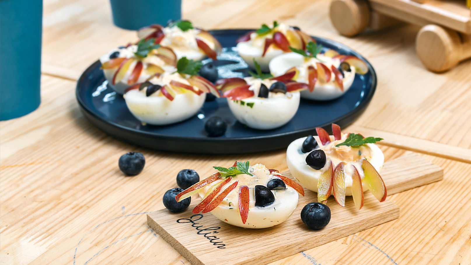 Auf einem Holztisch sind kreative Snacks angerichtet: kleine gekochte Eierhälften mit Äpfeln, Blaubeeren und frischen Kräutern dekoriert. Ein schwarzer Teller mit weiteren Snacks ist im Hintergrund sichtbar. Zudem sind einige Papierbecher und ein Holzspielzeug im Bild.