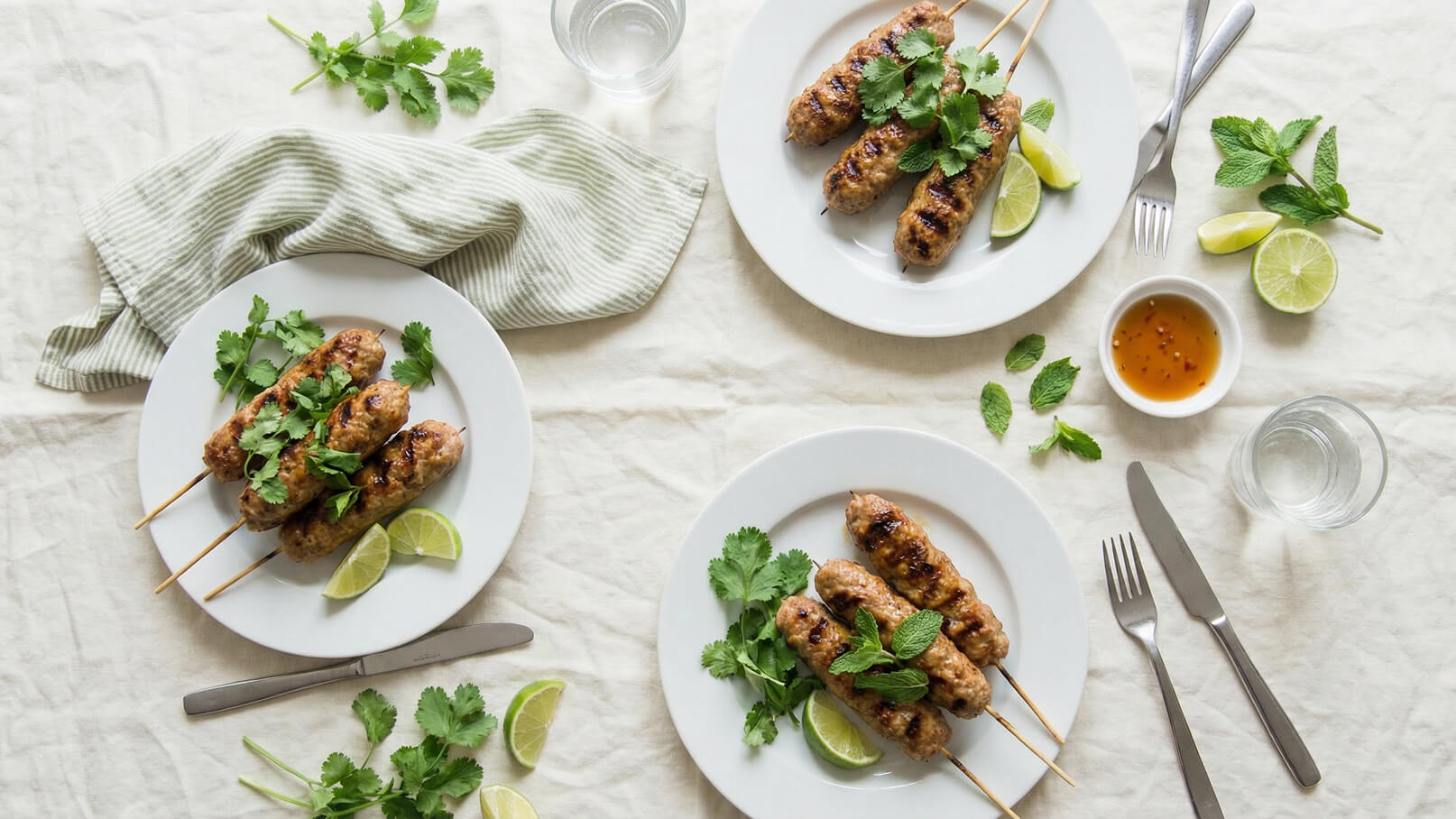 Auf einem Tisch liegen zwei weiße Teller mit gegrillten Fleischspießen, garniert mit frischem Koriander und Minze, sowie Limettenscheiben. Daneben steht eine kleine Schüssel mit einer süßlichen Sauce und ein Glas Wasser. Der Tisch ist mit einer hellen Tischdecke gedeckt.
