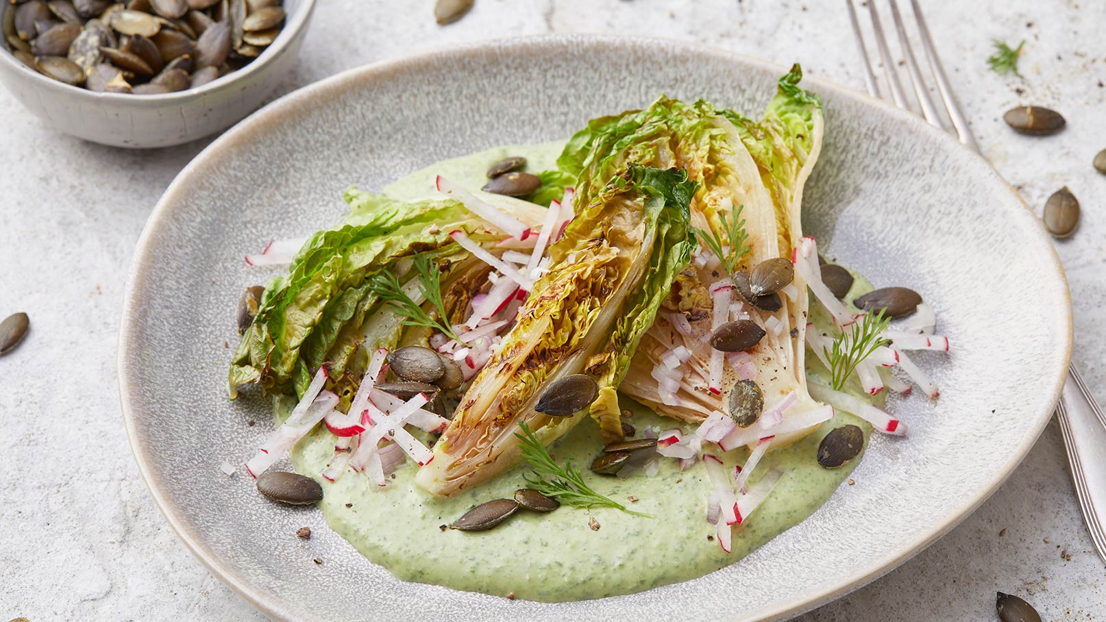 Ein Teller mit gegrilltem Romana-Salat, garniert mit Radieschenstreifen und Kürbiskernen, liegt auf einem hellen Untergrund. Der Salat befindet sich auf einer cremigen, grünen Sauce. Der Anblick vermittelt eine frische und gesunde Mahlzeit.