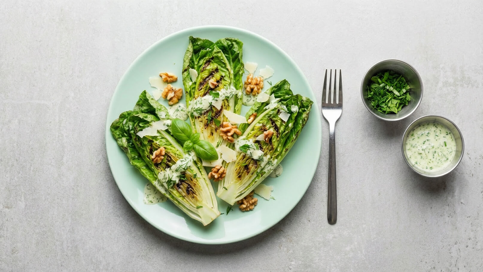 Ein grüner Salat auf einem mintfarbenen Teller, bestehend aus gegrilltem Romana-Salat, garniert mit Walnüssen, frischen Kräutern und Parmesan. Neben dem Teller befinden sich zwei kleine Schalen: eine mit gehackten Kräutern und eine mit einer Joghurtsauce. Ein Besteck liegt ebenfalls daneben.
