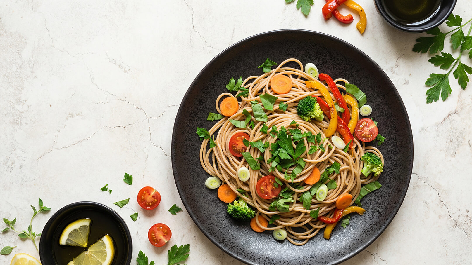 Ein schwarzer Teller mit Spaghetti, garniert mit frischem Gemüse wie Cherrytomaten, Karotten, Brokkoli und Paprika. Darüber liegen gehackte Kräuter, die dem Gericht Farbe verleihen. Neben dem Teller befinden sich eine kleine Schüssel mit Olivenöl und einige Zitronenscheiben.