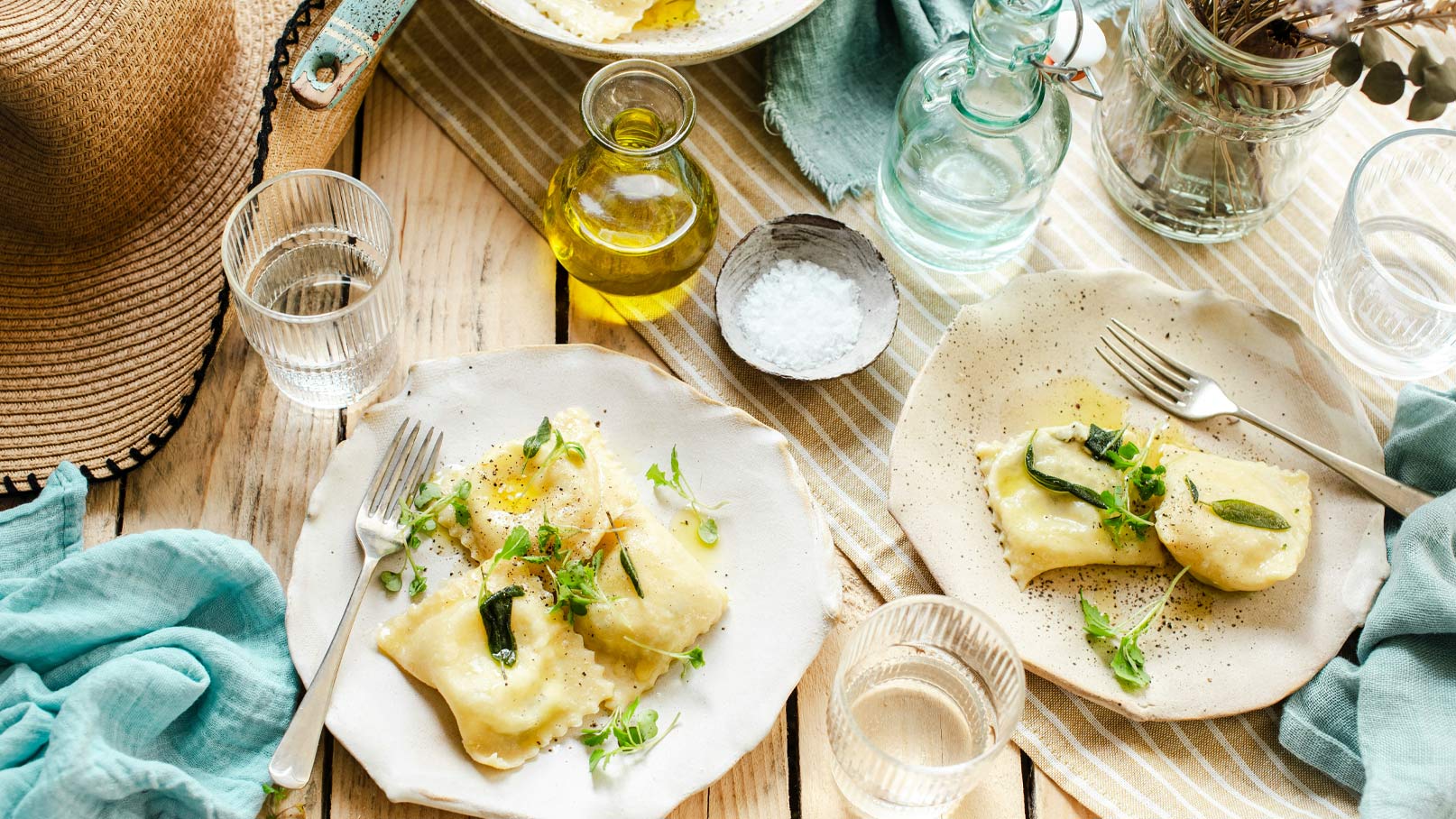 Auf dem Tisch stehen zwei Teller mit frischen Ravioli, garniert mit Kräutern und einem Spritzer Öl. Eine Gabel liegt neben einem der Teller. Weitere Utensilien wie Gläser und eine Flasche Öl sind ebenfalls sichtbar, sowie ein gestreiftes Tuch.