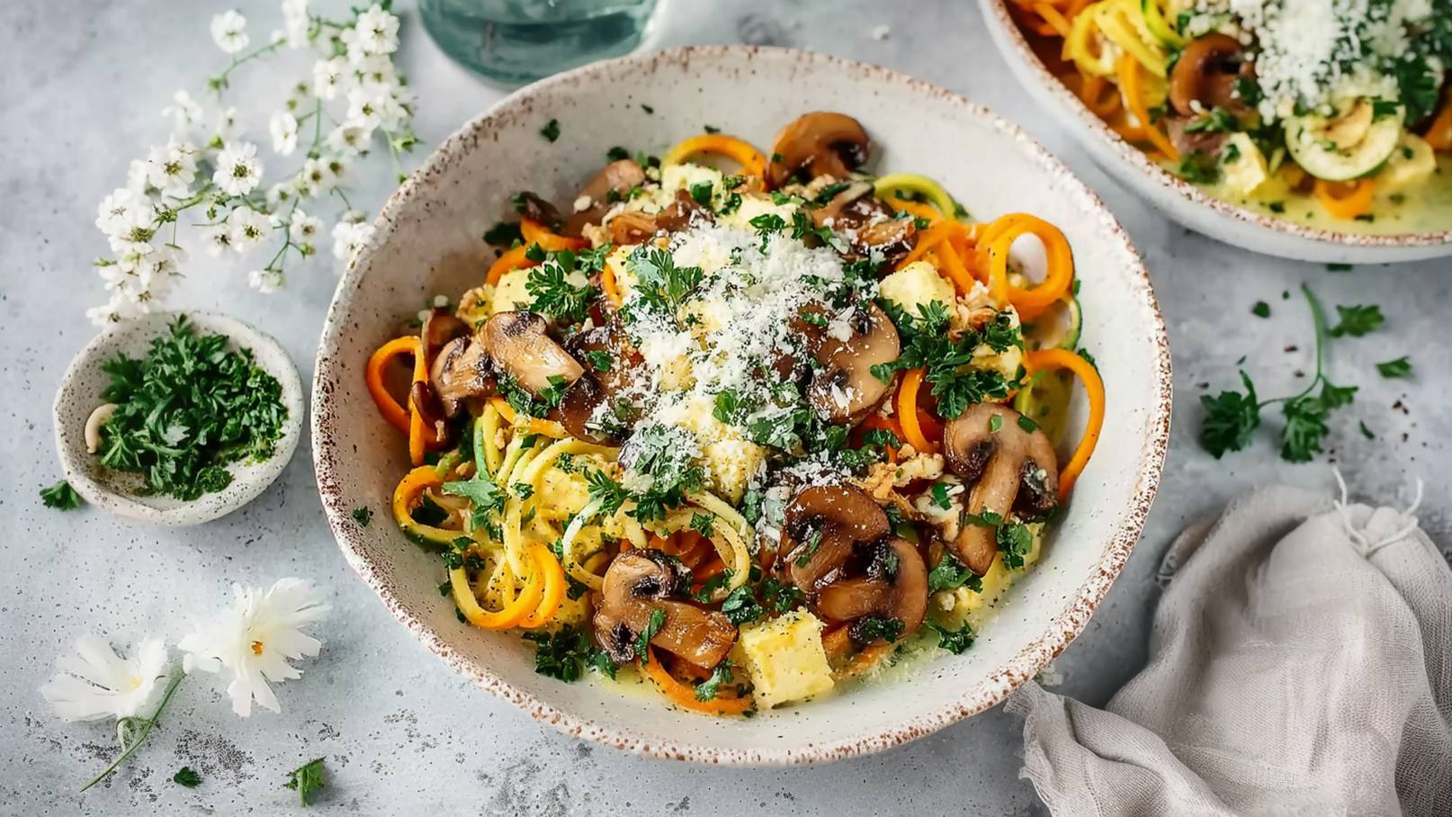 Eine Schüssel mit bunten Gemüsenudeln, darunter Karotten- und Zucchini-Spaghetti, garniert mit gebratenen Champignons und frischen Kräutern. Darauf verteilt liegt geriebener Parmesan. Im Hintergrund sind ein kleiner Schälchen mit Kräutern und Besenblumen zu sehen.