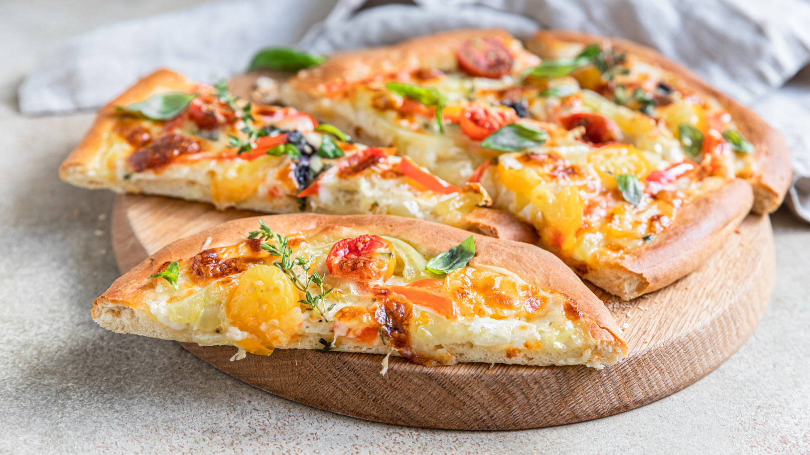 Drei Stücke Pizza liegen auf einem runden Holzbrett. Die Pizza ist mit geschmolzenem Käse, buntem Gemüse und frischen Kräutern belegt, darunter Tomaten und Paprika. Die Kruste ist goldbraun und leicht knusprig. Eine Portion wurde bereits herausgeschnitten.