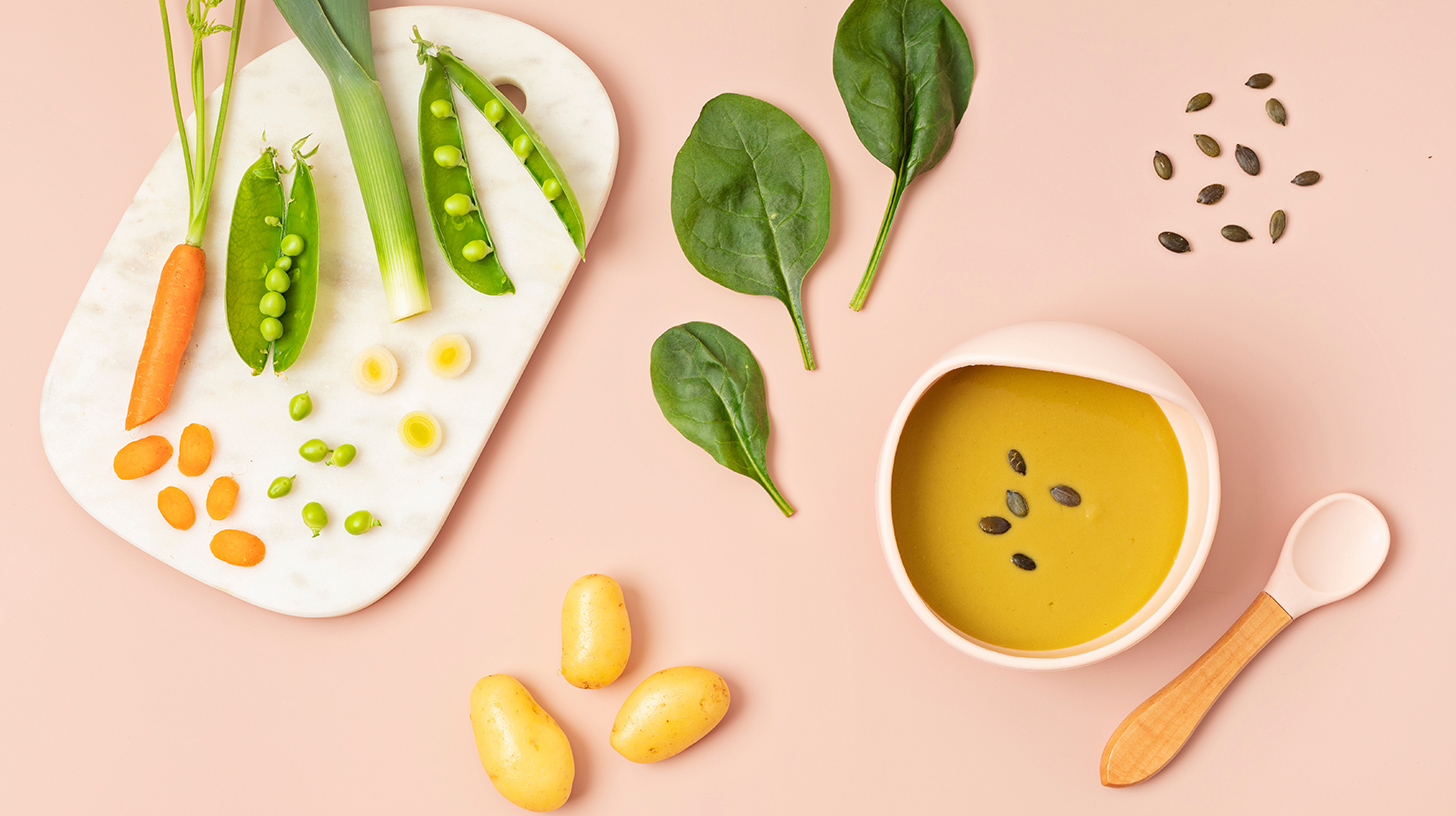 Eine helle, cremige Suppe in einer ovalen Schüssel, garniert mit Kürbiskernen. Um die Schüssel herum liegen frische Zutaten: eine Karotte, Lauch, frische Spinatblätter, Erbsen in der Schote und kleine Kartoffeln auf einem weißen Schneidebrett. Der Hintergrund ist rosa.