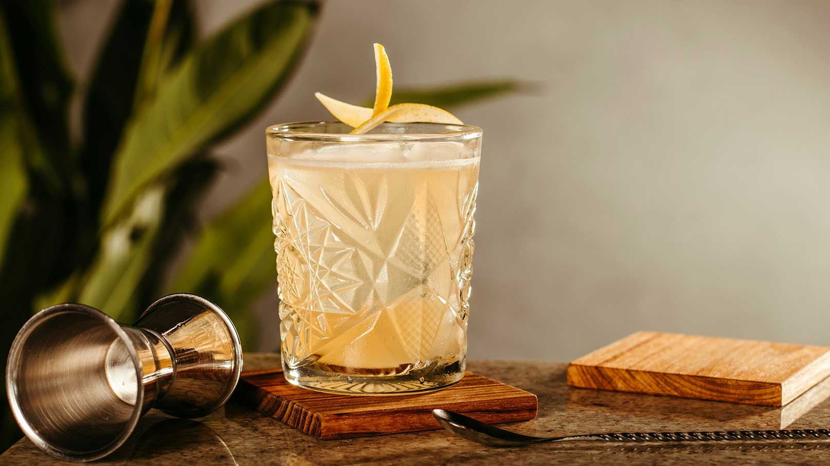 Ein Glas mit einem hellen Cocktail steht auf einem Holzuntersetzer, dekoriert mit einer Zitronenscheibe. Daneben liegt ein Silbermessbecher und ein Holzblock. Im Hintergrund sind unscharfe grüne Pflanzenblätter zu sehen.