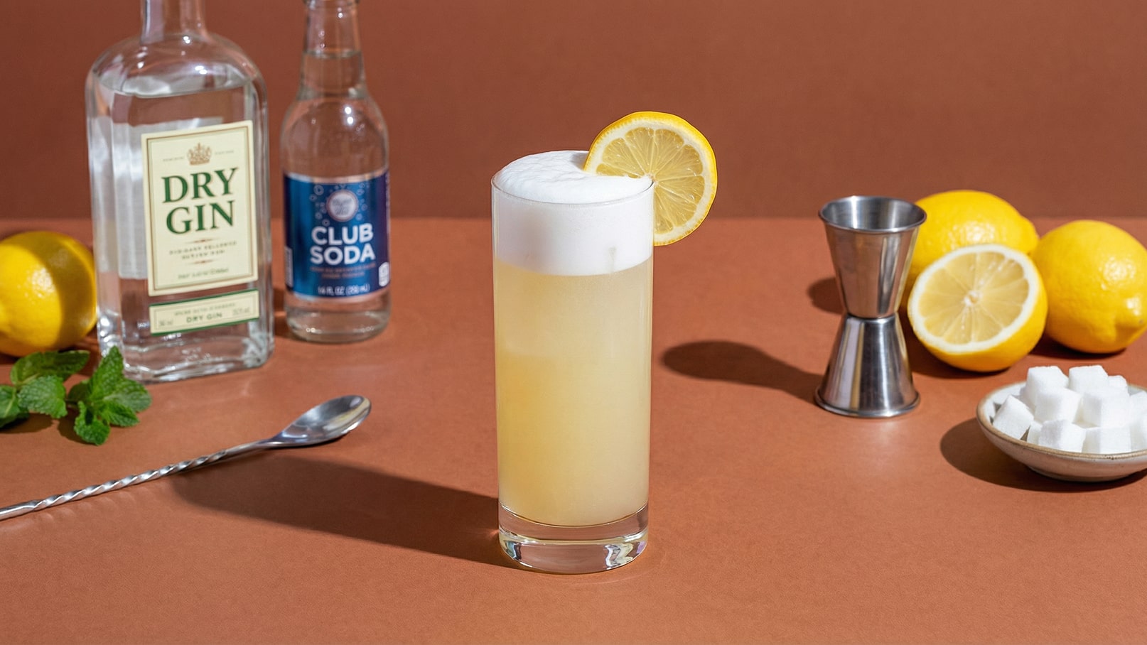 Ein hoher Cocktailglas mit schäumendem, zitronenfarbenem Getränk und einer Zitronenscheibe am Rand. Daneben stehen eine Flasche Dry Gin und ein Club Soda. Im Hintergrund sind weitere Zitronen und Zuckerwürfel auf einem Teller sichtbar.