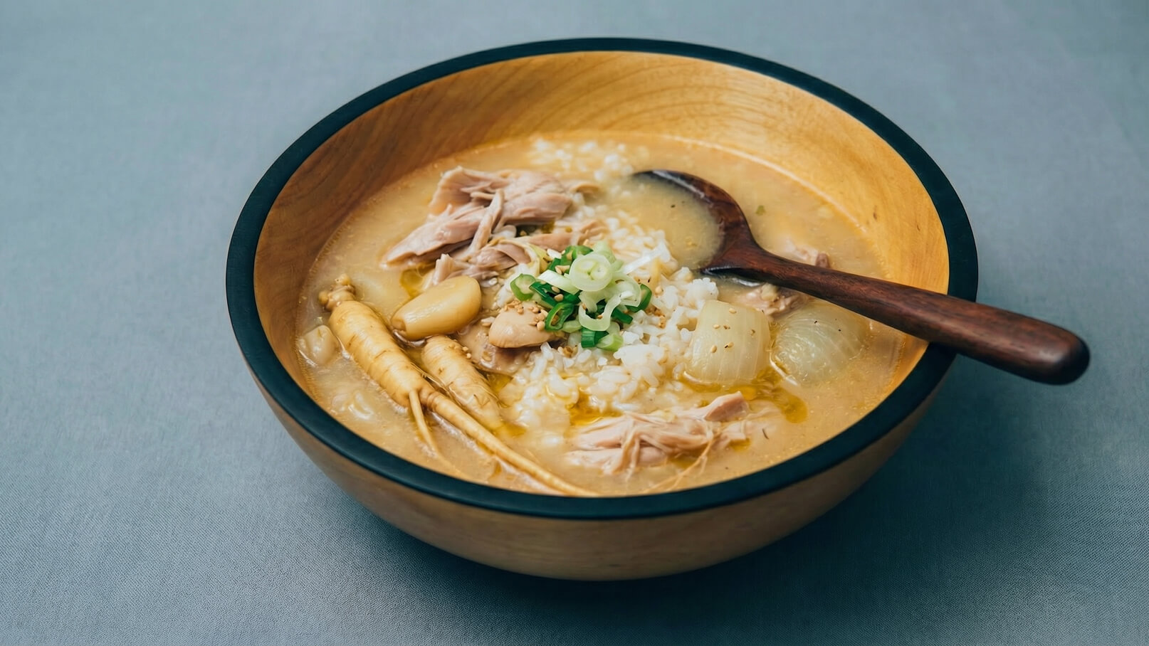 Eine Holzschüssel enthält eine schmackhafte Suppe mit Hühnerfleisch, Reis, Gemüse und Kräutern. Ein Holzlöffel liegt neben der Suppe. Der Hintergrund ist grau und unauffällig, um den Fokus auf das Gericht zu lenken.
