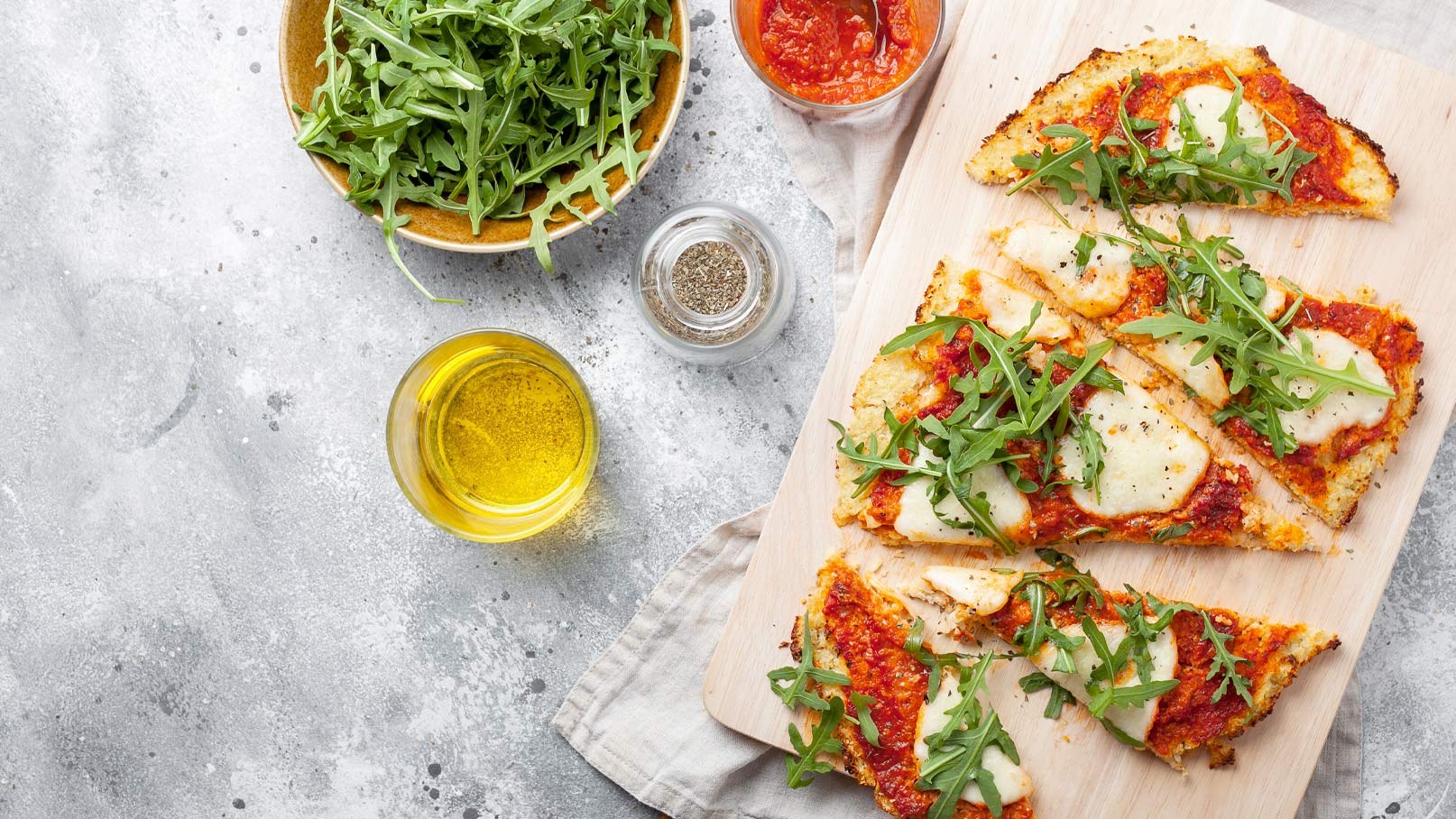 Ein Holzbrett mit mehreren Pizzastücken, belegt mit Tomatensoße, Käse und frischem Rucola. Daneben steht eine Schale mit Rucola, ein Glas mit einer gelben Flüssigkeit und ein Glas mit roter Sauce. Ein Gewürzstreuer ist ebenfalls zu sehen.